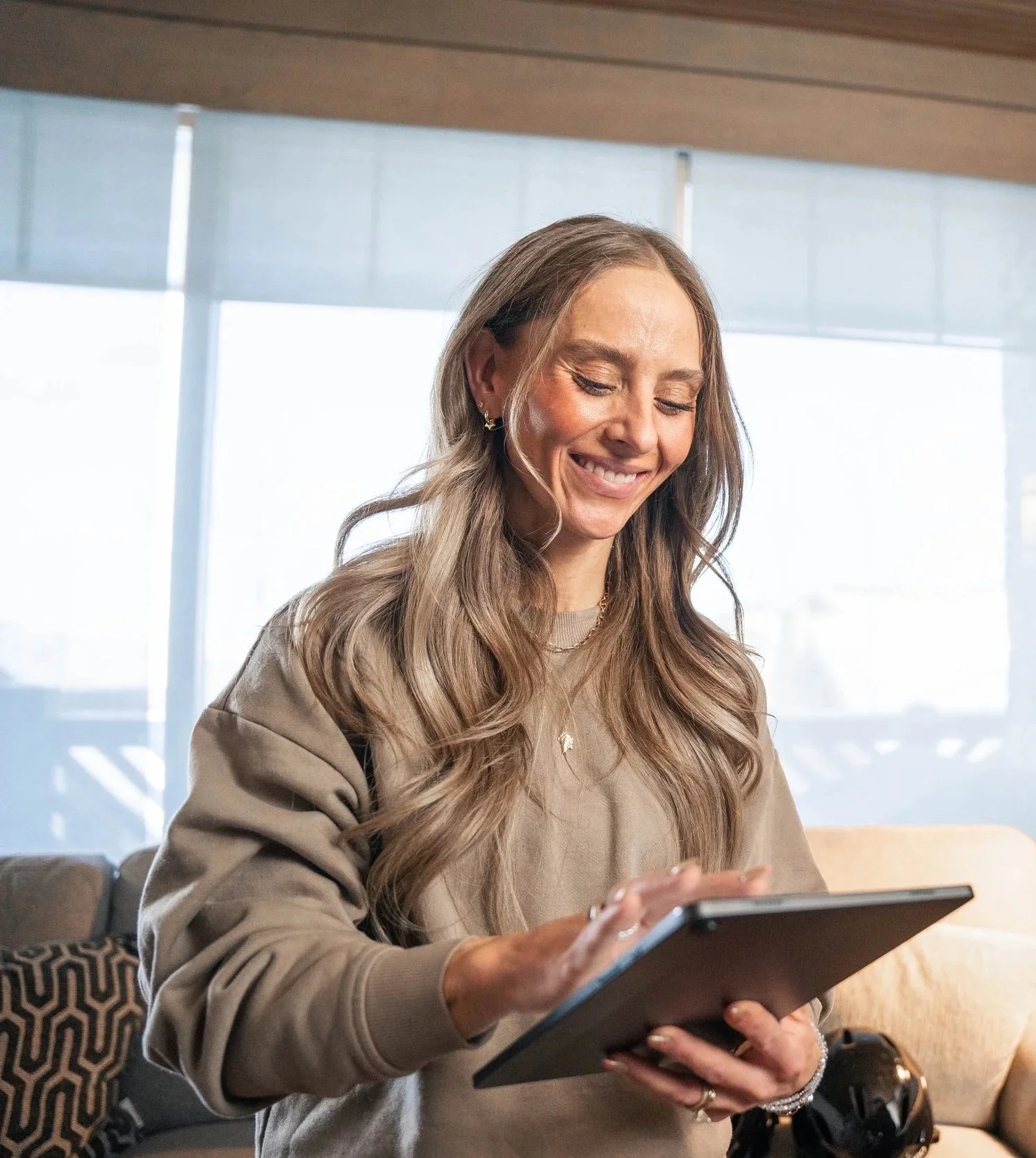 A woman with long wavy hair smiling while holding a tablet in a bright indoor setting.