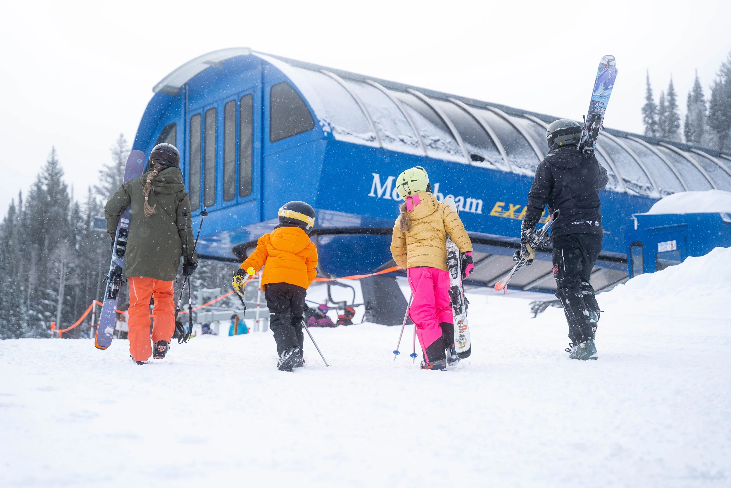Children and adults in winter clothing holding skis, preparing to ski near a blue ski lift station in a snowy mountain landscape.