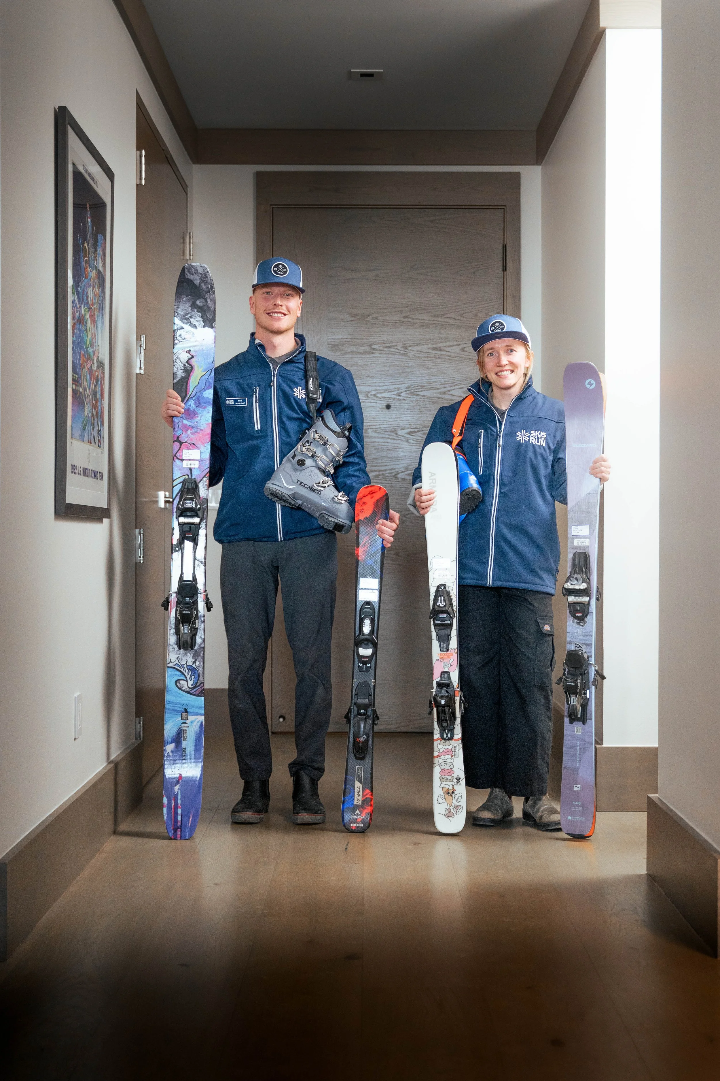 Two people, a man and a woman, standing indoors holding skis and ski boots, smiling at the camera. They are wearing dark blue jackets and matching baseball caps with a logo, and the background features a wooden door and a framed poster on the wall.