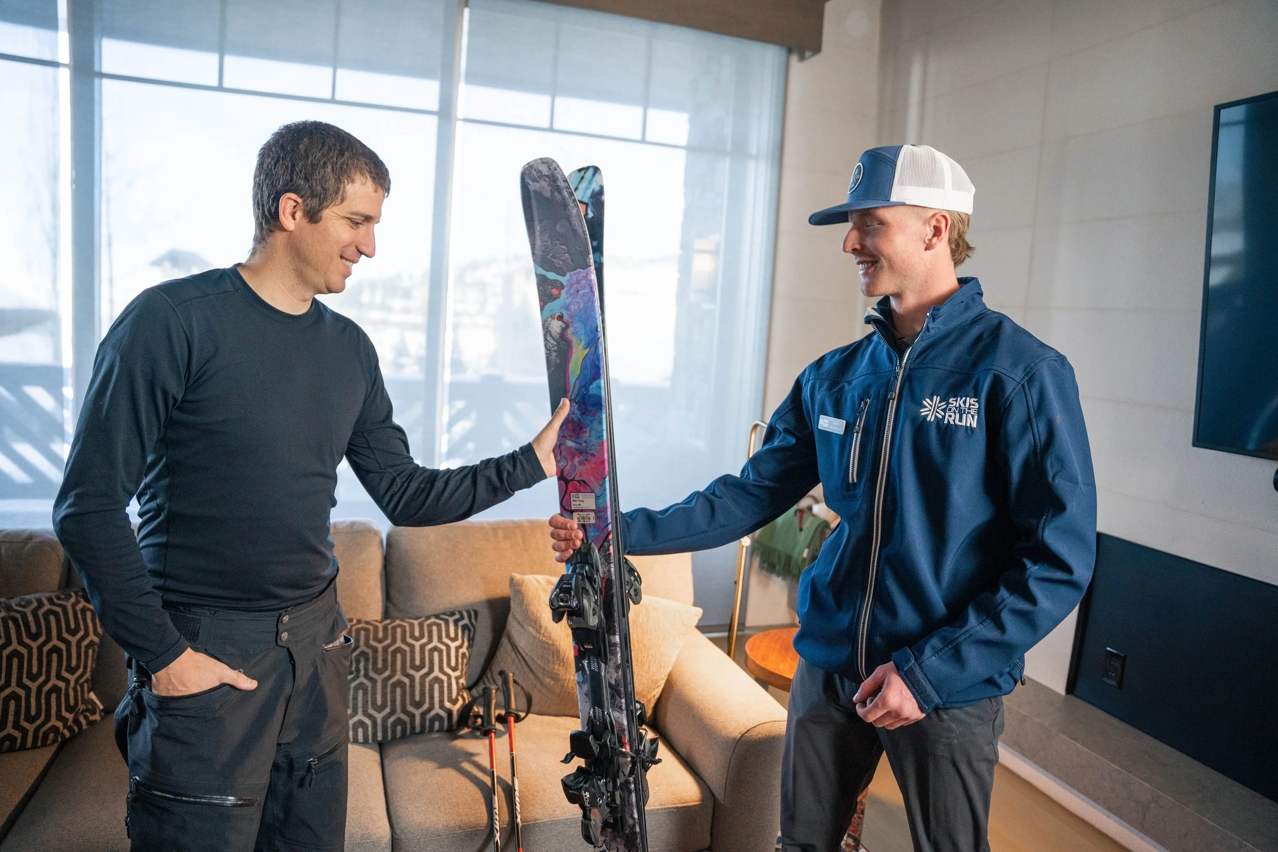 Two men are indoors, one is holding a colorful pair of skis while the other is smiling and pointing at them. The man with skis is wearing a blue jacket and a baseball cap, while the other is dressed in a black long sleeve shirt. Behind them are large windows and a sofa.