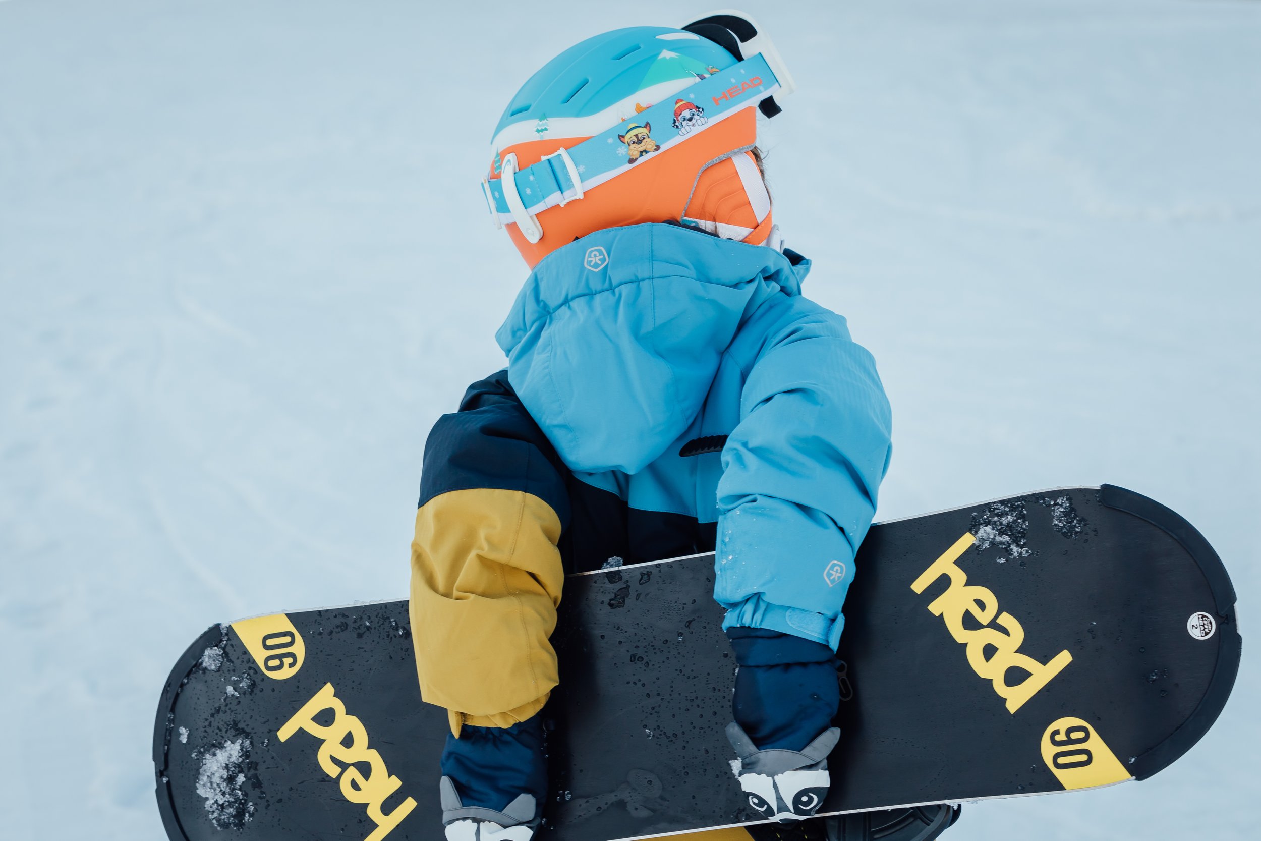A person wearing a blue helmet and a colorful ski jacket, holding a snowboard, standing on snow.