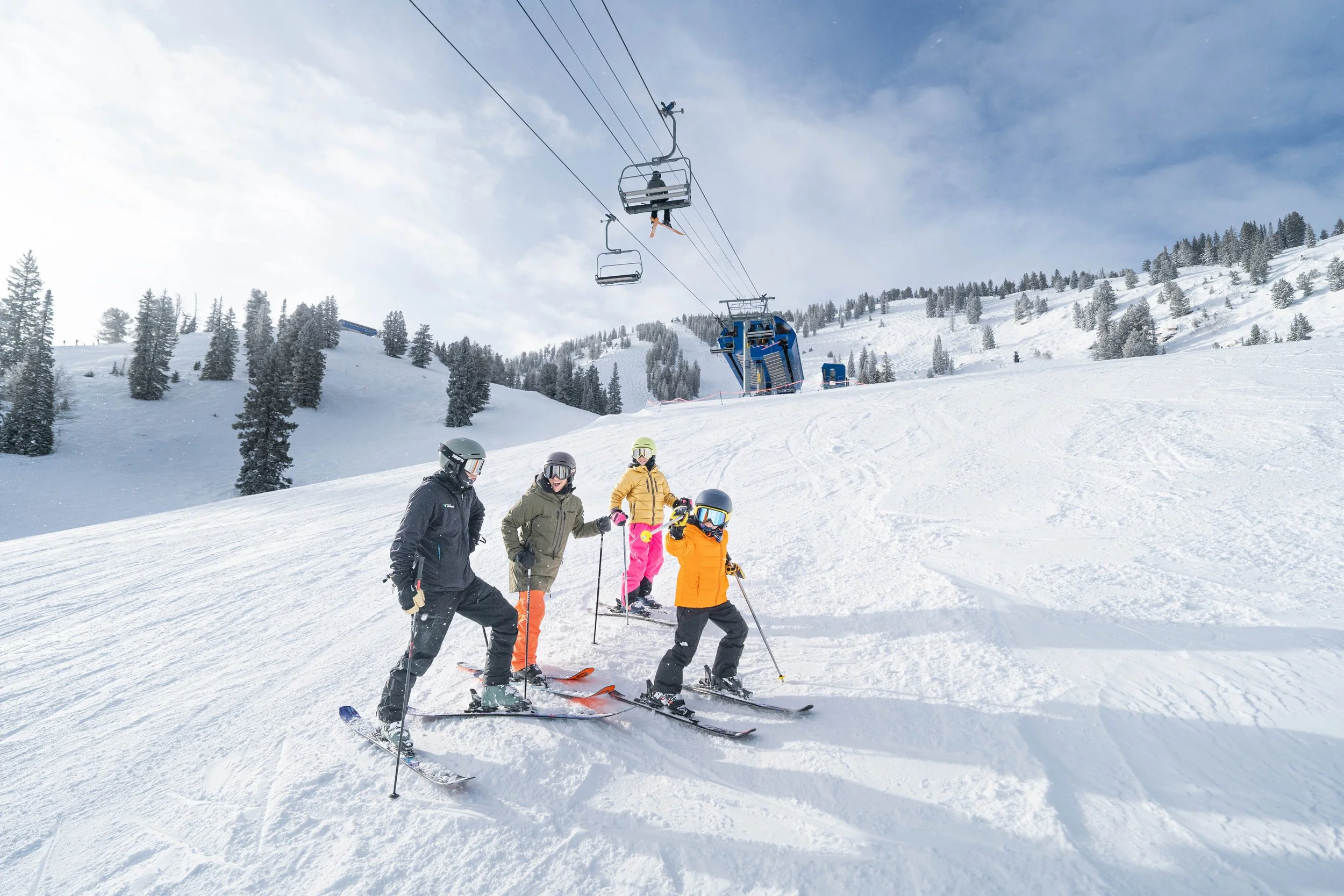 A group of four people in ski gear standing on snow, with ski poles, in front of a snowy mountain landscape, while two ski lifts are visible in the background.