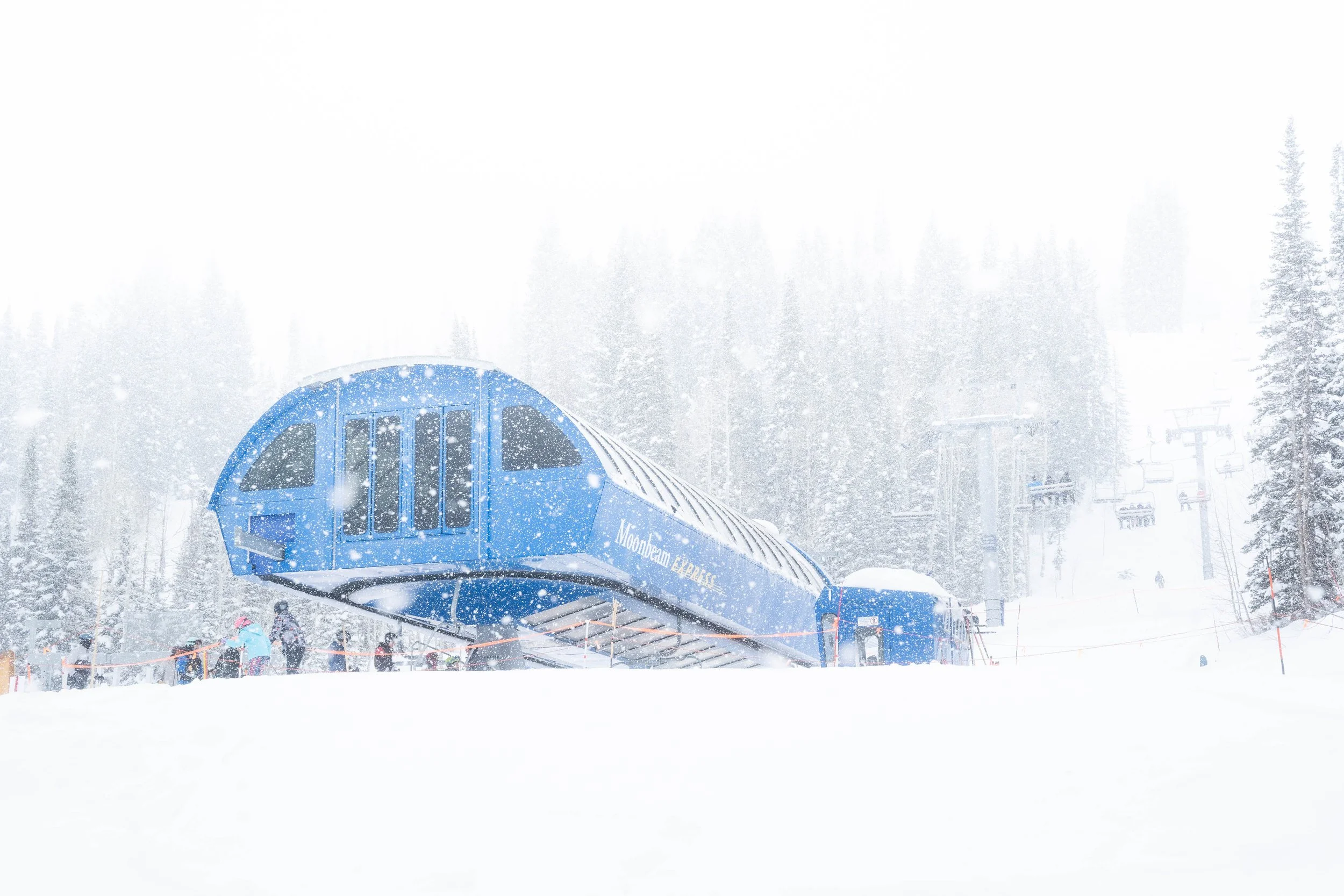 A ski lift station in a snowy mountain landscape with snow falling and ski slopes, trees, and skiers visible in the background.