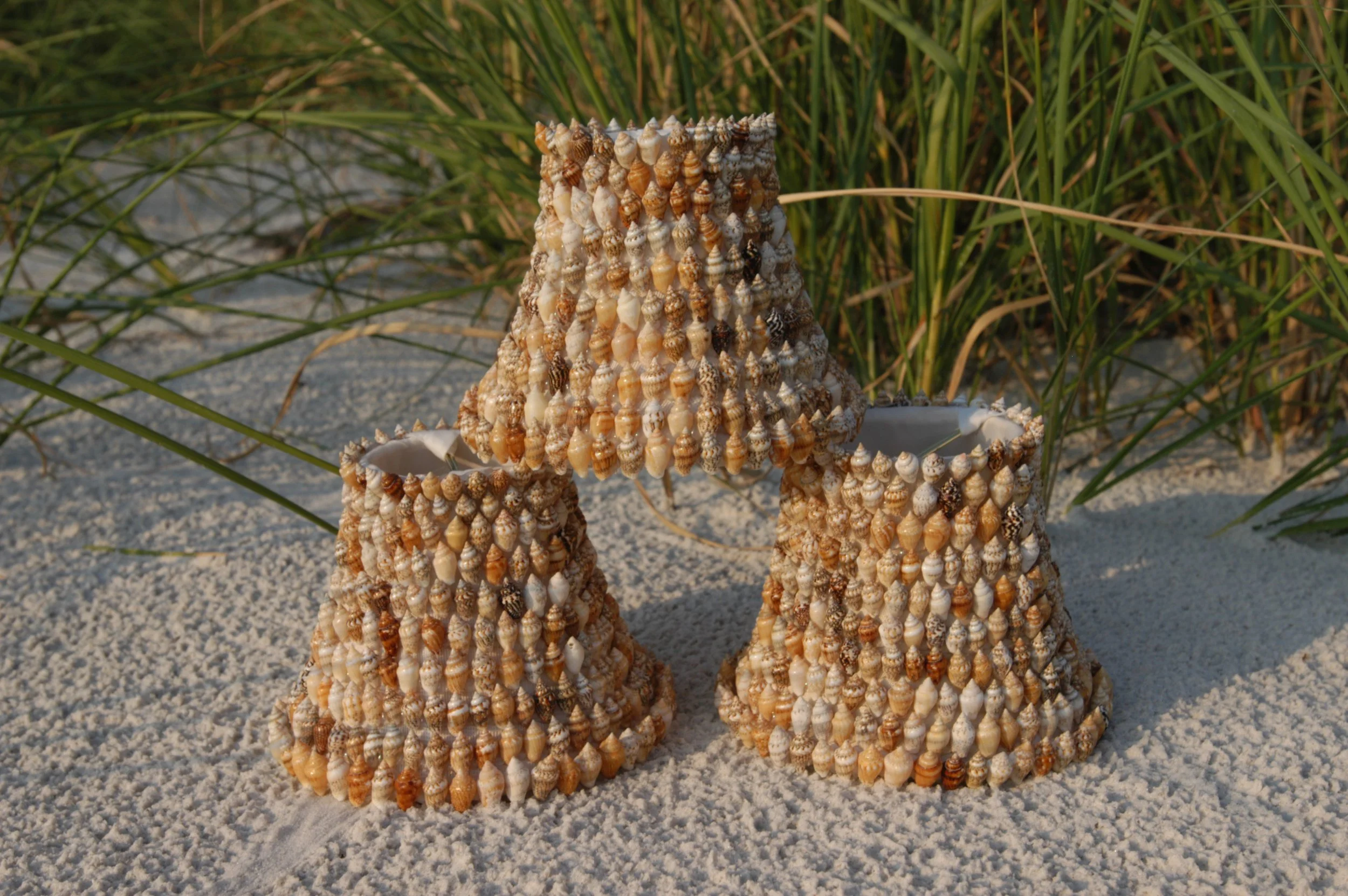 Three cone-shaped sculptures covered with small seashells on a sandy beach with green grass in the background.