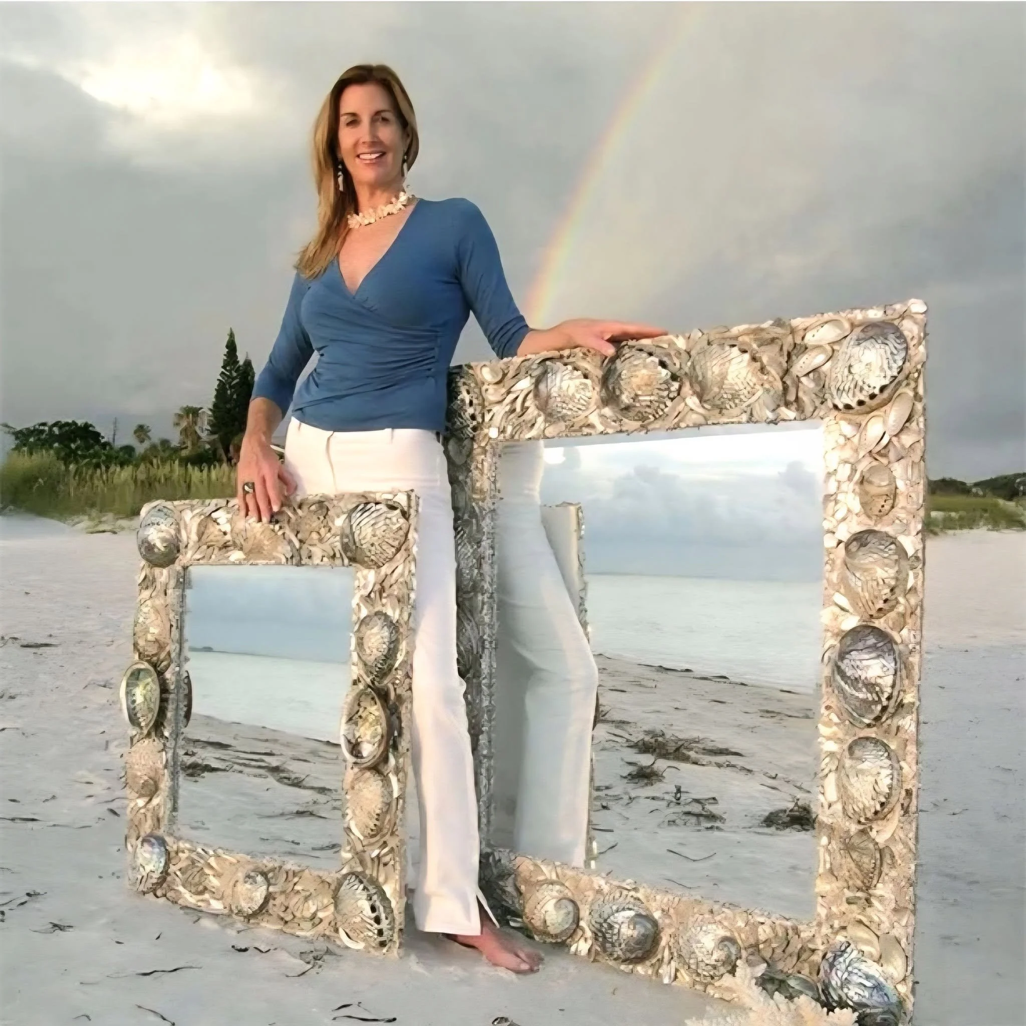 A woman in a blue top and white pants standing outdoors at sunset, smiling with a rainbow in the cloudy sky behind her, near a structure made of seashells.