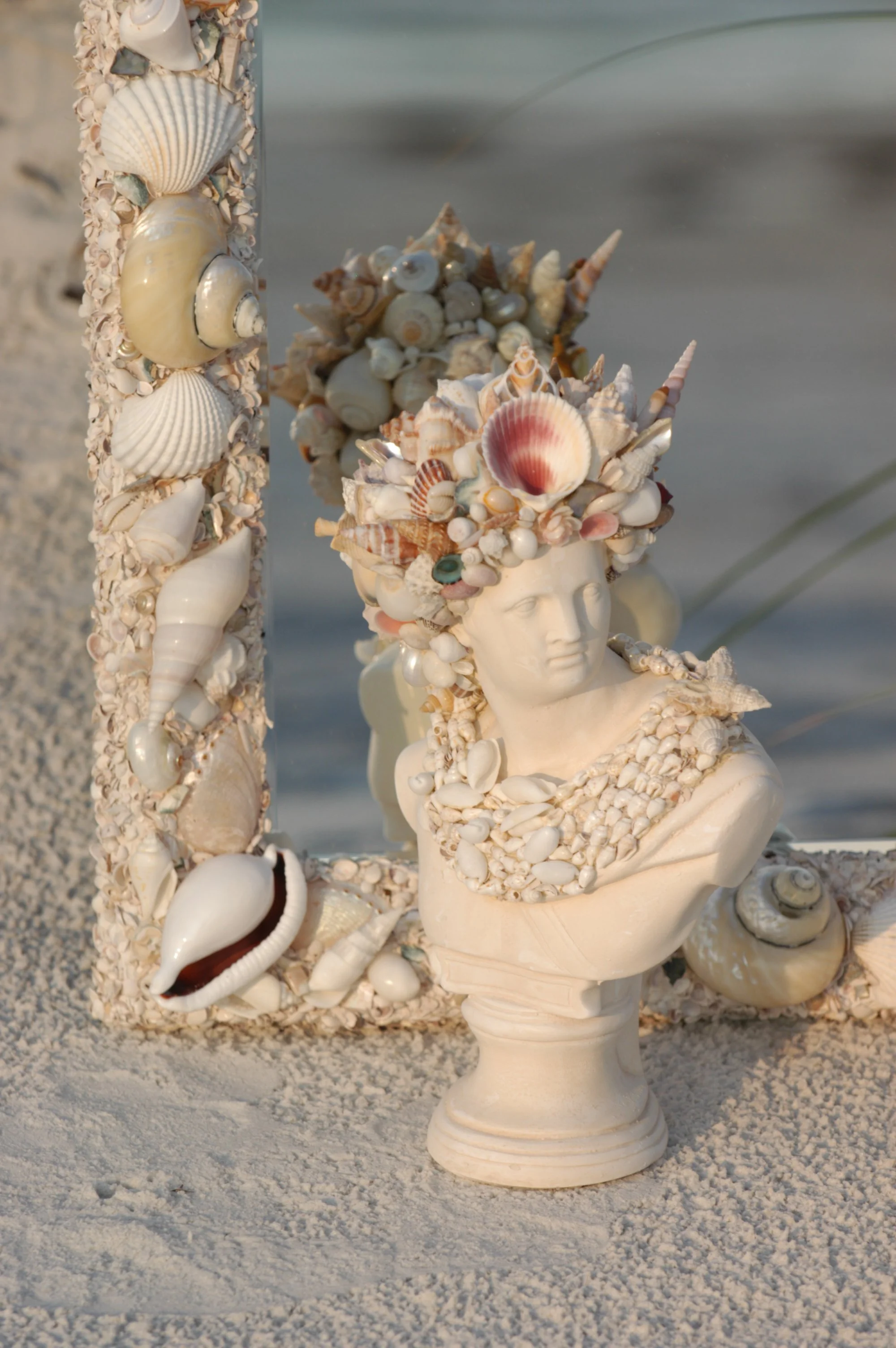 A bust sculpture of a woman adorned with a beautiful seashell crown and shell jewelry, displayed on a sandy surface, sitting in front of a large seashell mirror adorned with expensive hand selected seashells.
