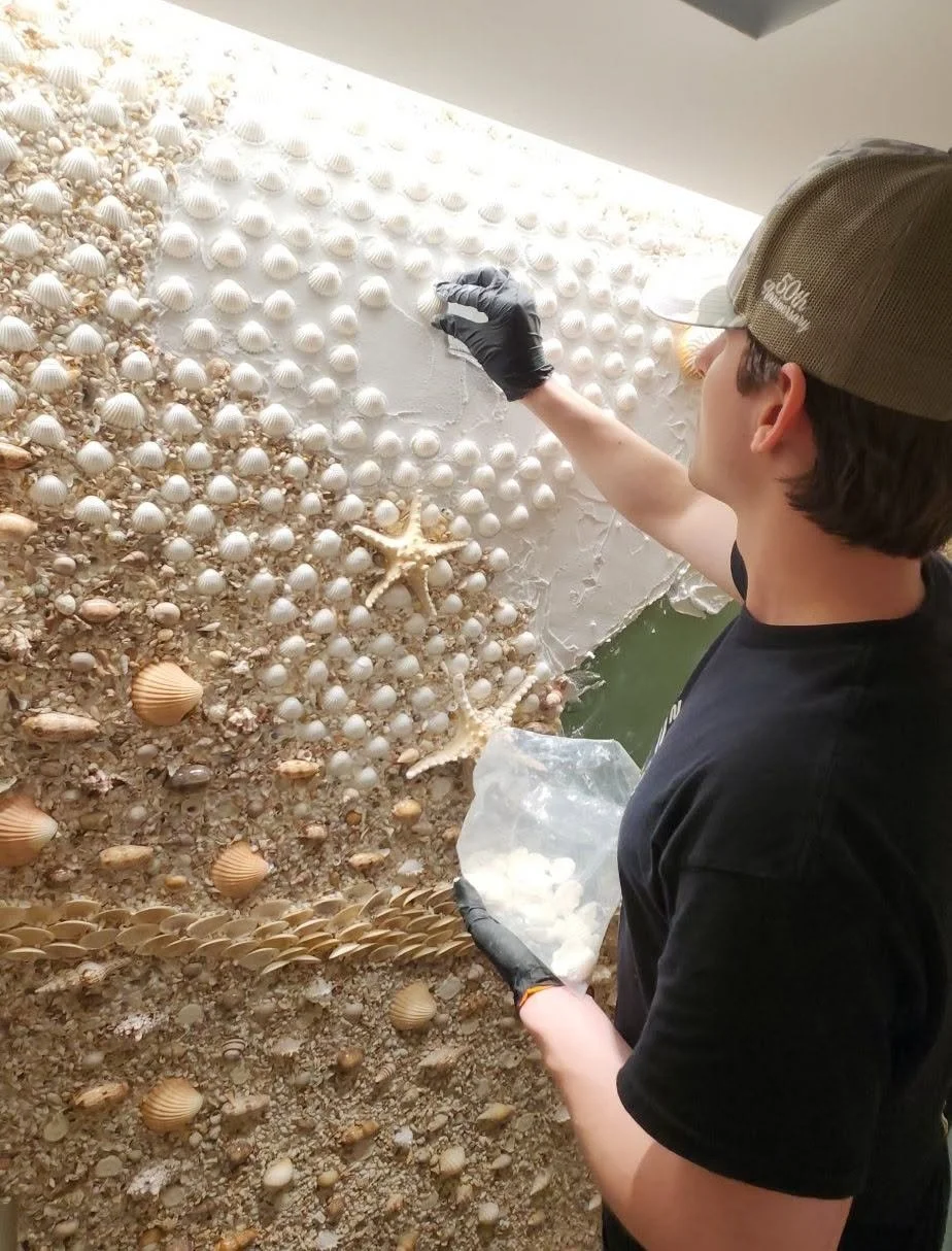 A person wearing a tan cap and black gloves is affixing small seashells and starfish to a wall, creating a decorative mosaic beach scene.