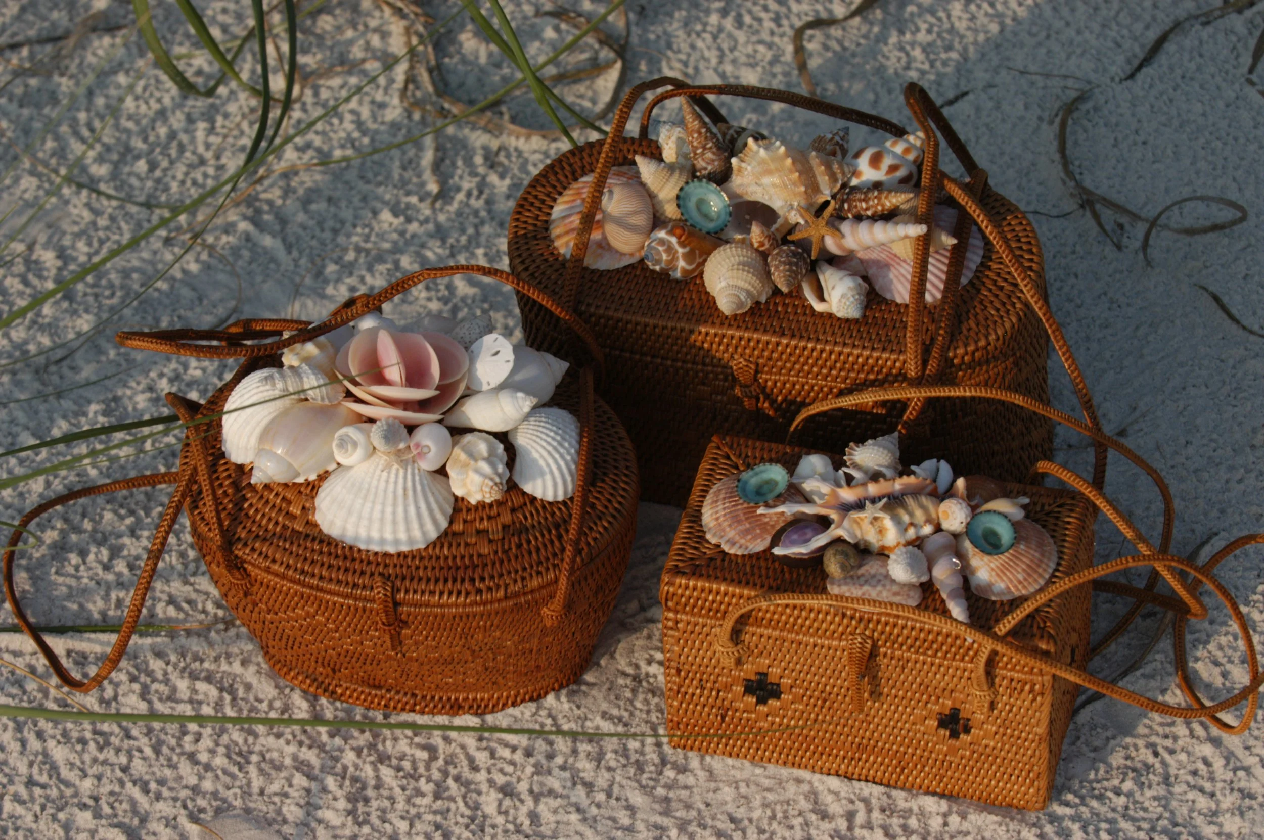 Three woven baskets filled with various seashells, placed on white sandy beach with green grass blades nearby.