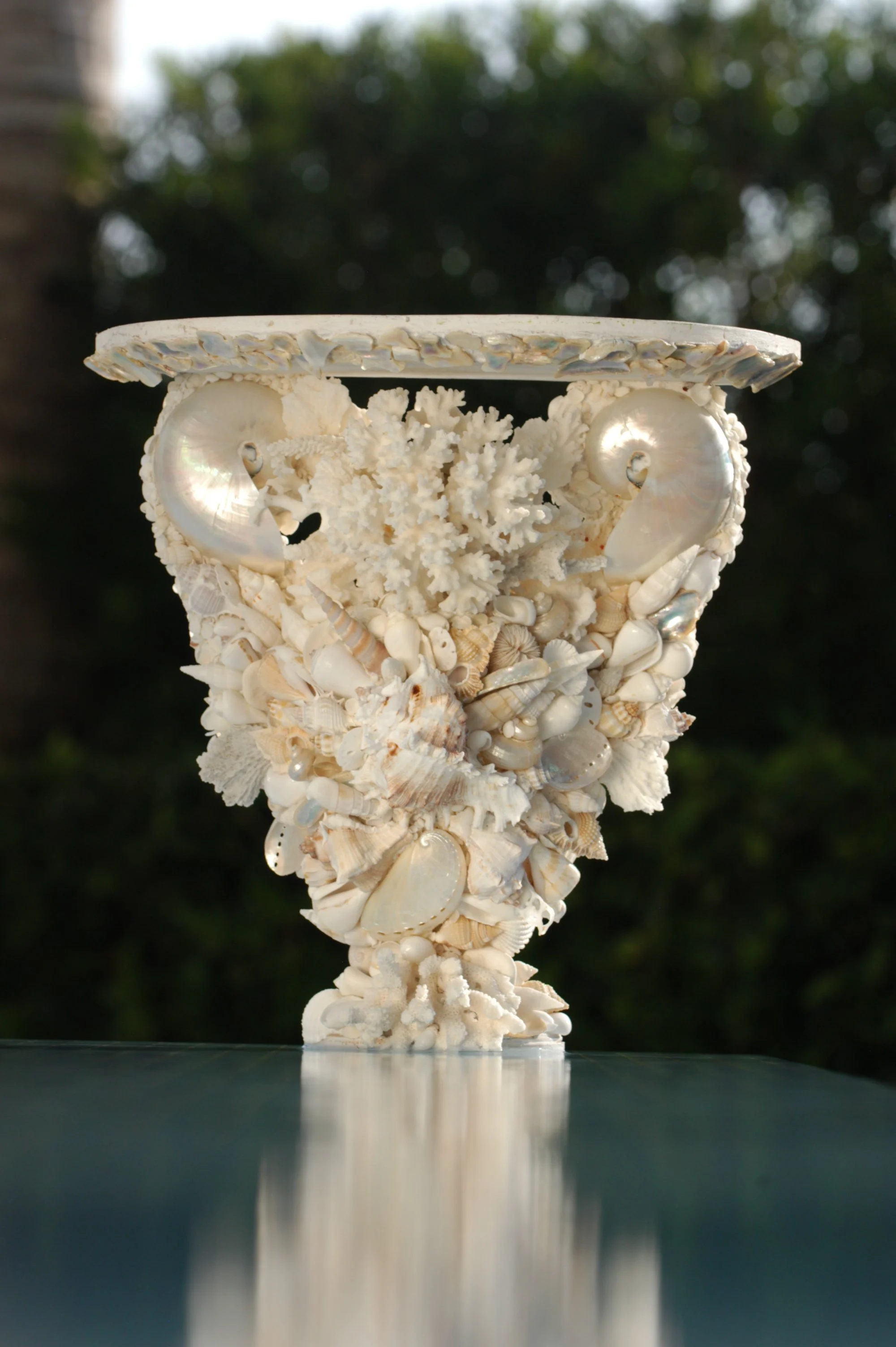 A tall sculpture made of polished seashells, coral, and other marine shells arranged on a pedestal, with a blurred outdoor background of trees.