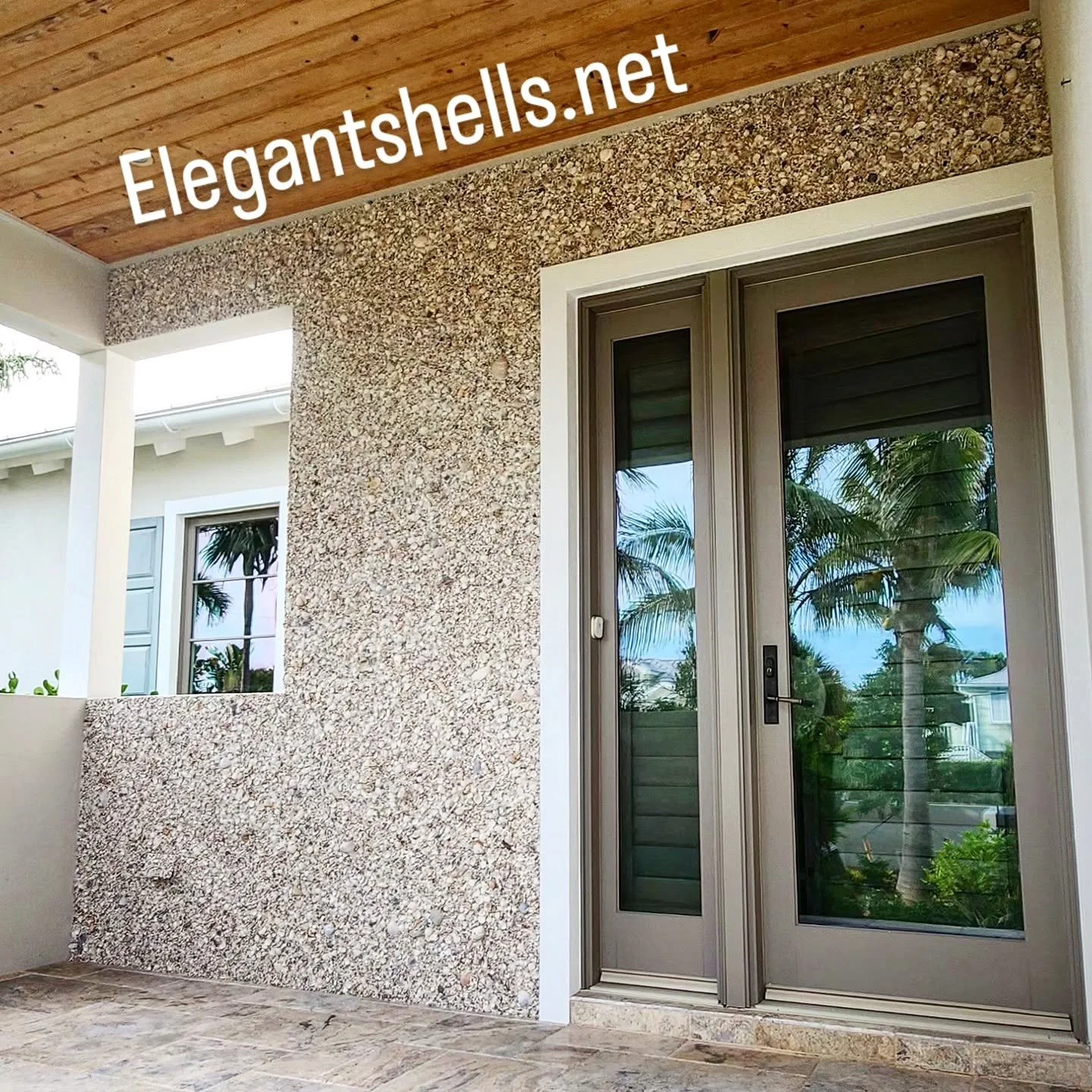 Exterior view of a house with sliding glass door, textured wall, and a wooden ceiling over a patio, with palm trees reflected in the glass.