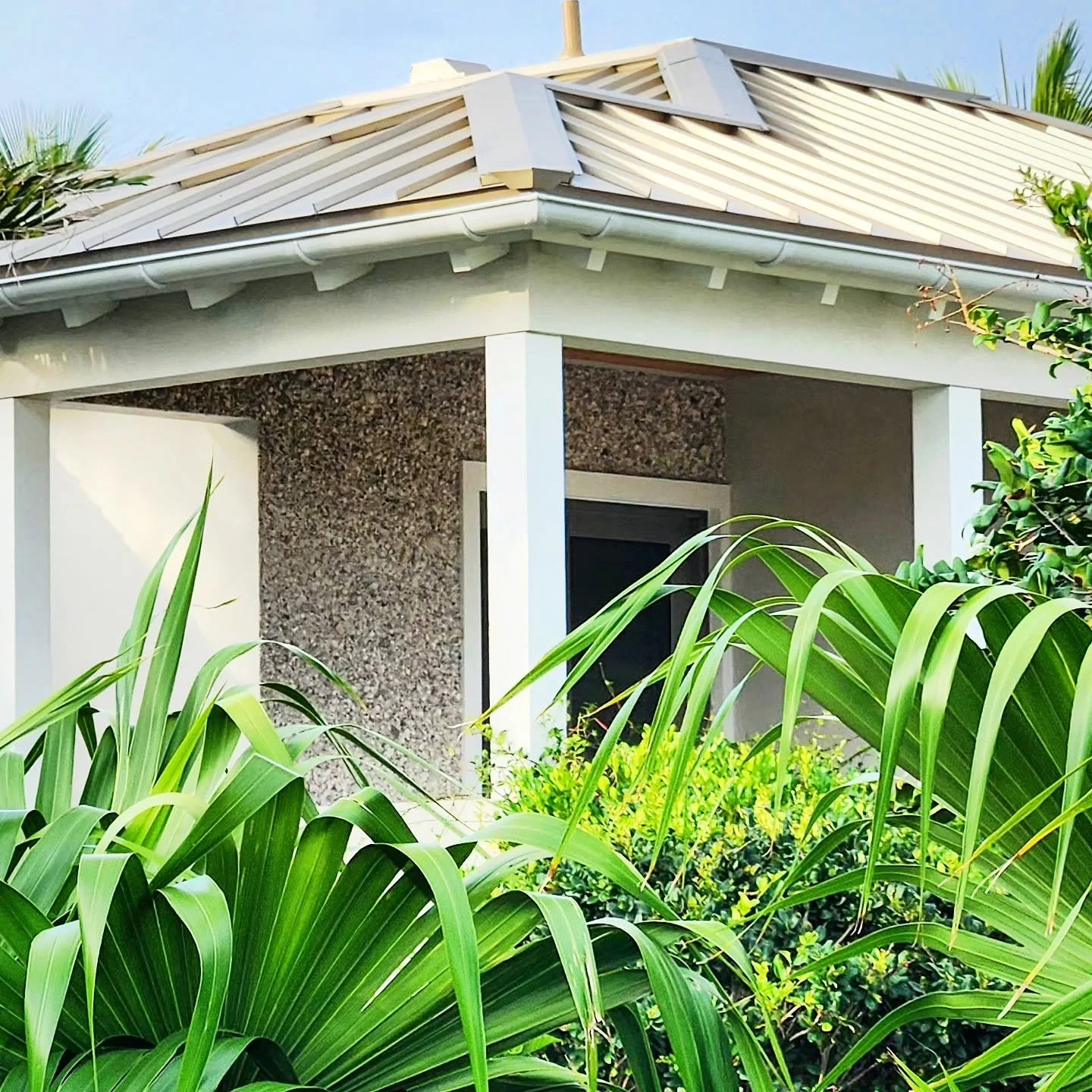 Part of a house with a beige metal roof, white support beams, a textured wall, and lush green tropical plants in the foreground.