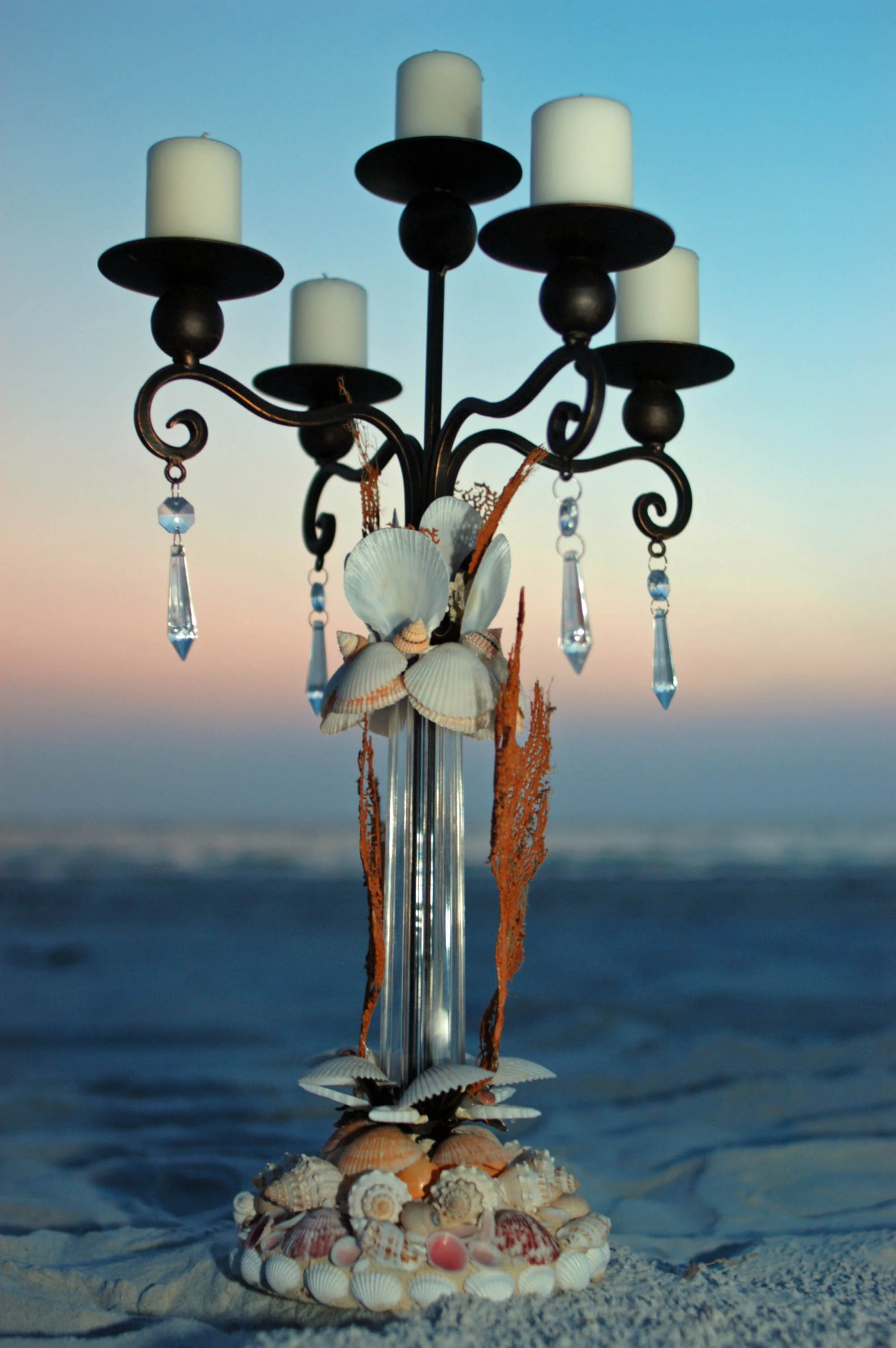 A beach scene with a decorative centerpiece made of seashells, glass beads, and candle holders, positioned on the sand with the ocean and sky in the background.