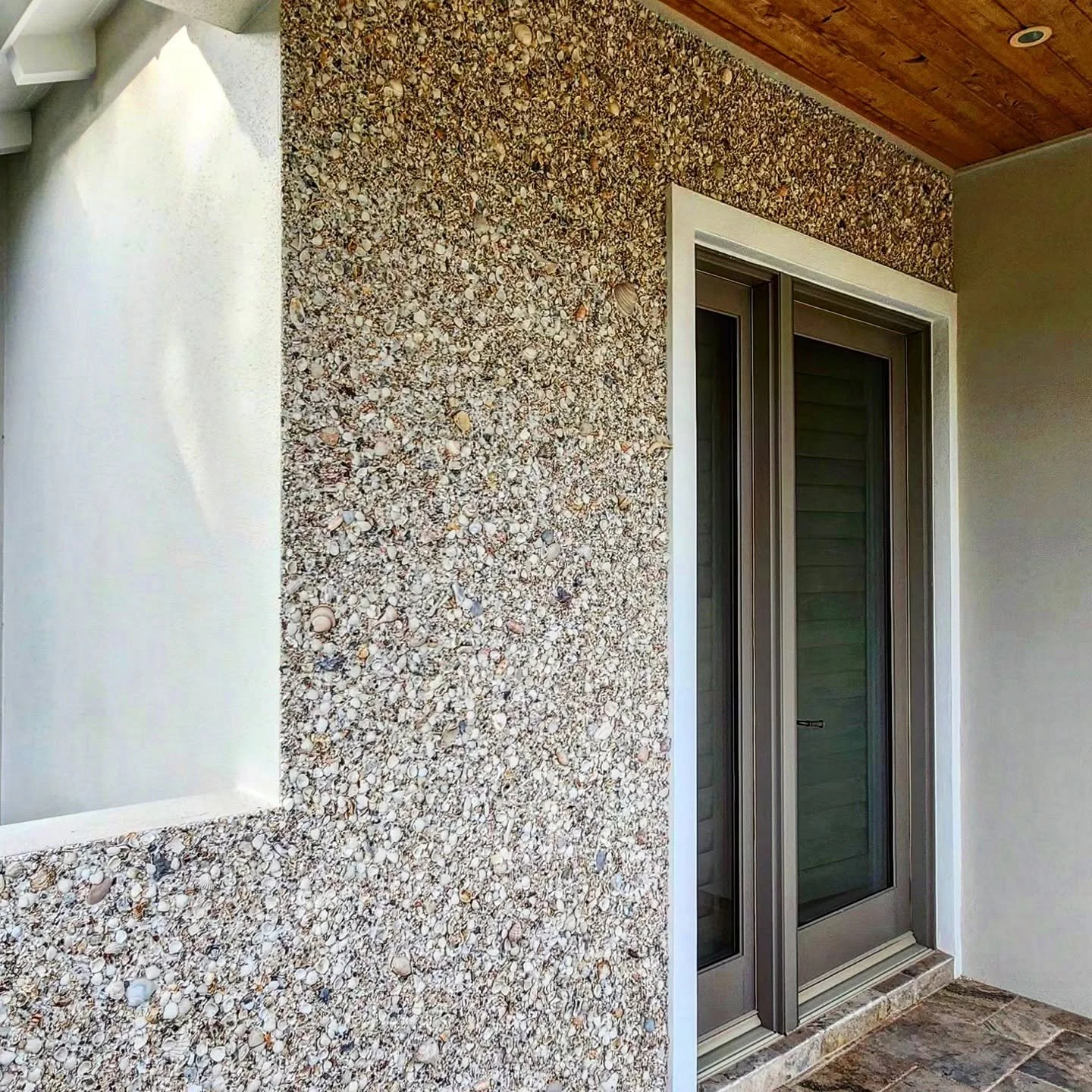 A corner of a room featuring a textured wall made of small seashells, a sliding glass door with a metal frame, a white window sill, and a wooden ceiling. The floor has tiles with a natural stone appearance.