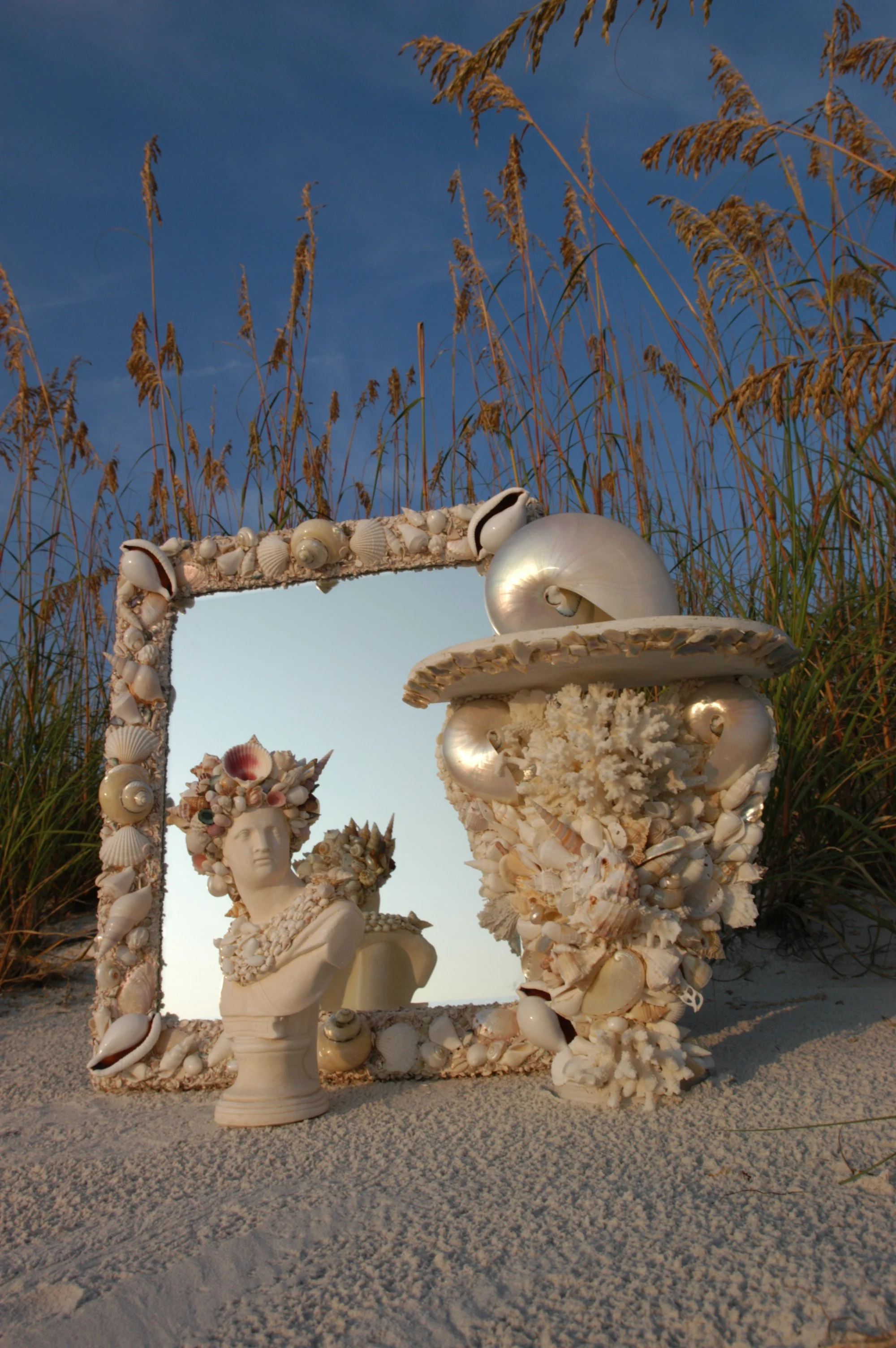 Coastal art installation made of white and iridescent seashells, featuring a bust sculpture, mirror, and a decorative shell arrangements, set outdoors on the beach with tall grasses in the background.