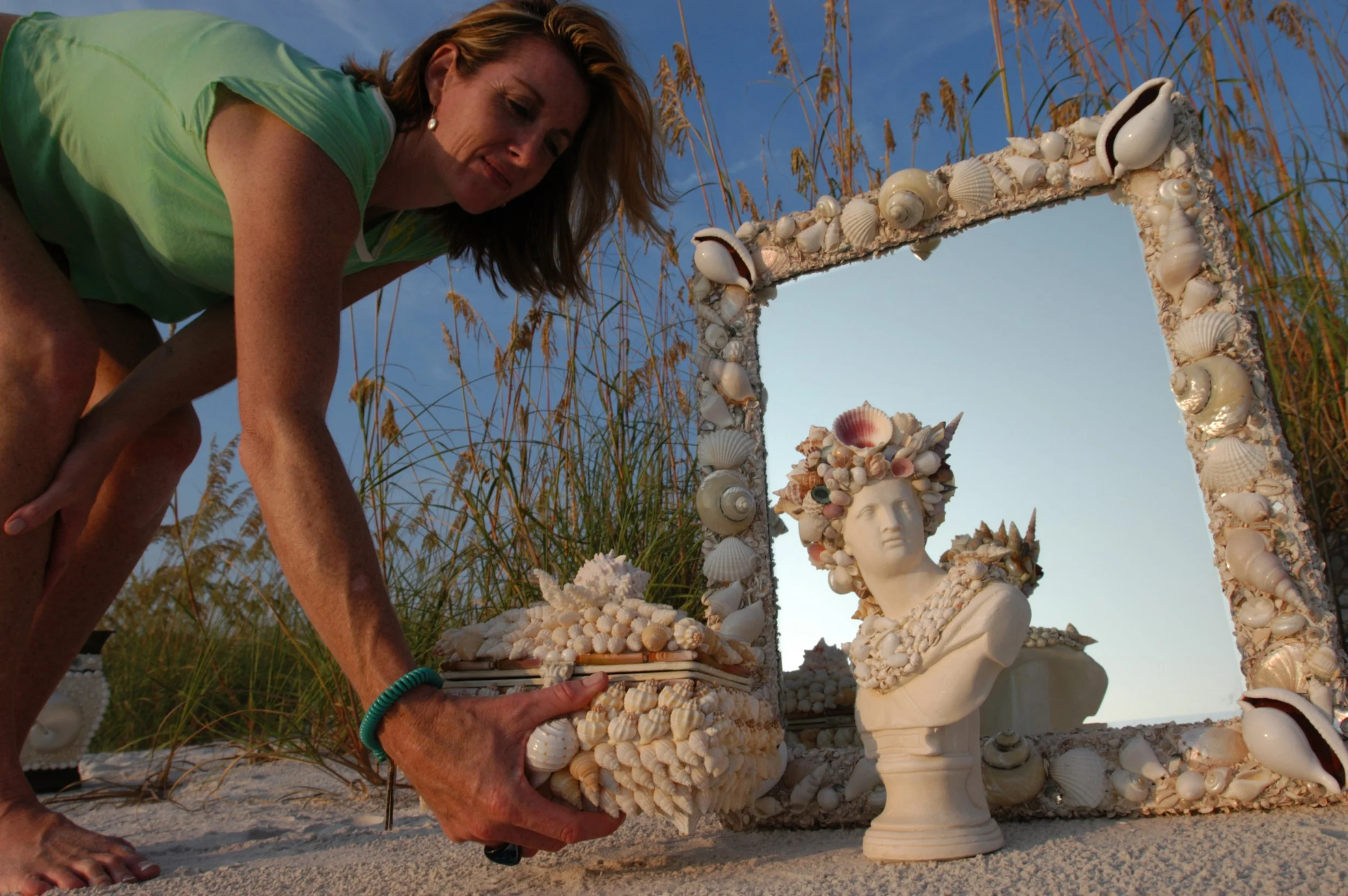 A woman places a seashell decorated jewelry box on the sand during sunset at the beach. In the background, there is a white seashell-embellished mirror and bust of a woman adorned with seashells.