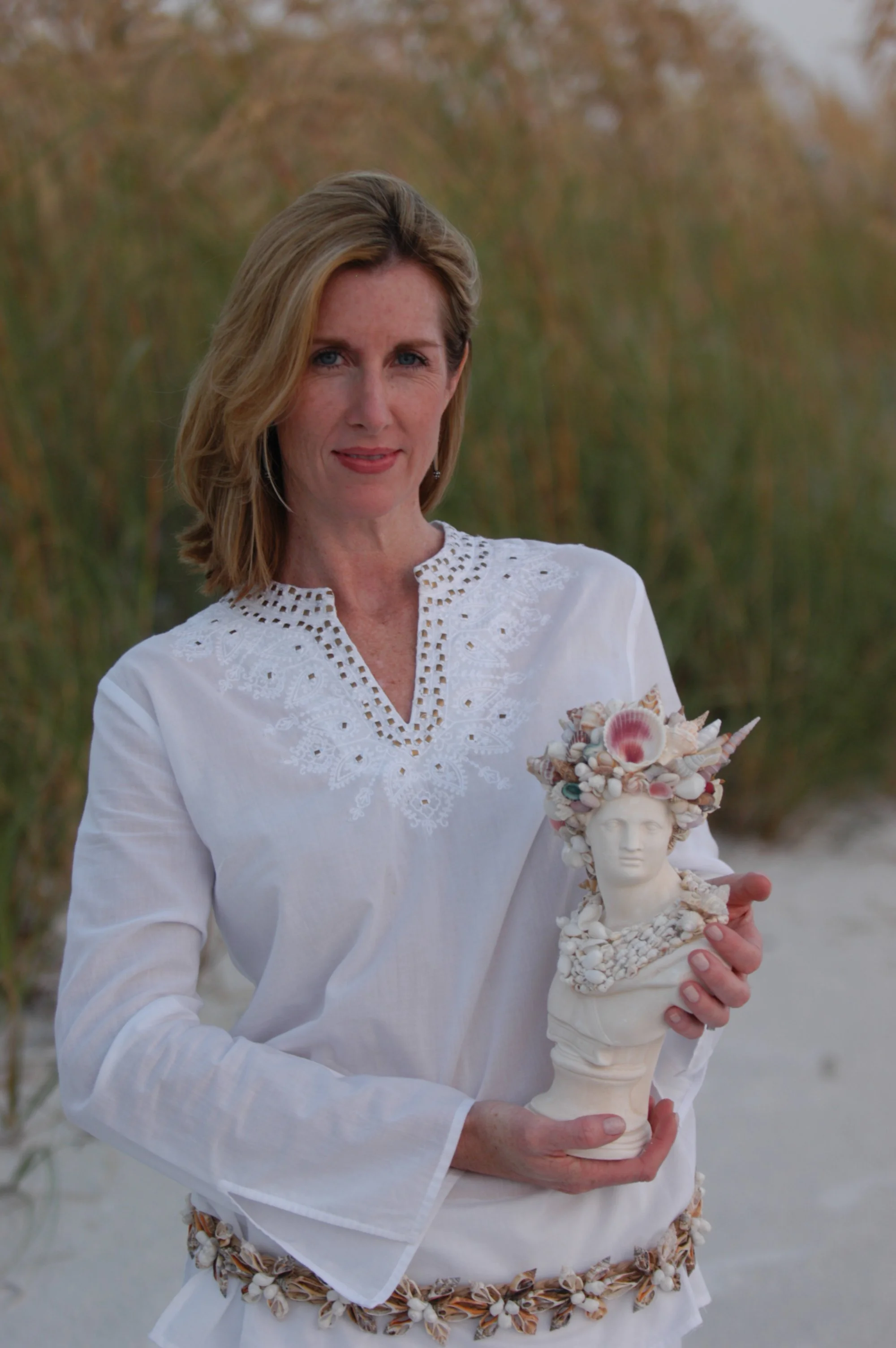A woman with blonde hair wearing a white embroidered blouse holds a decorative bust sculpture adorned with seashells and floral elements, standing outdoors with a blurred background of trees.