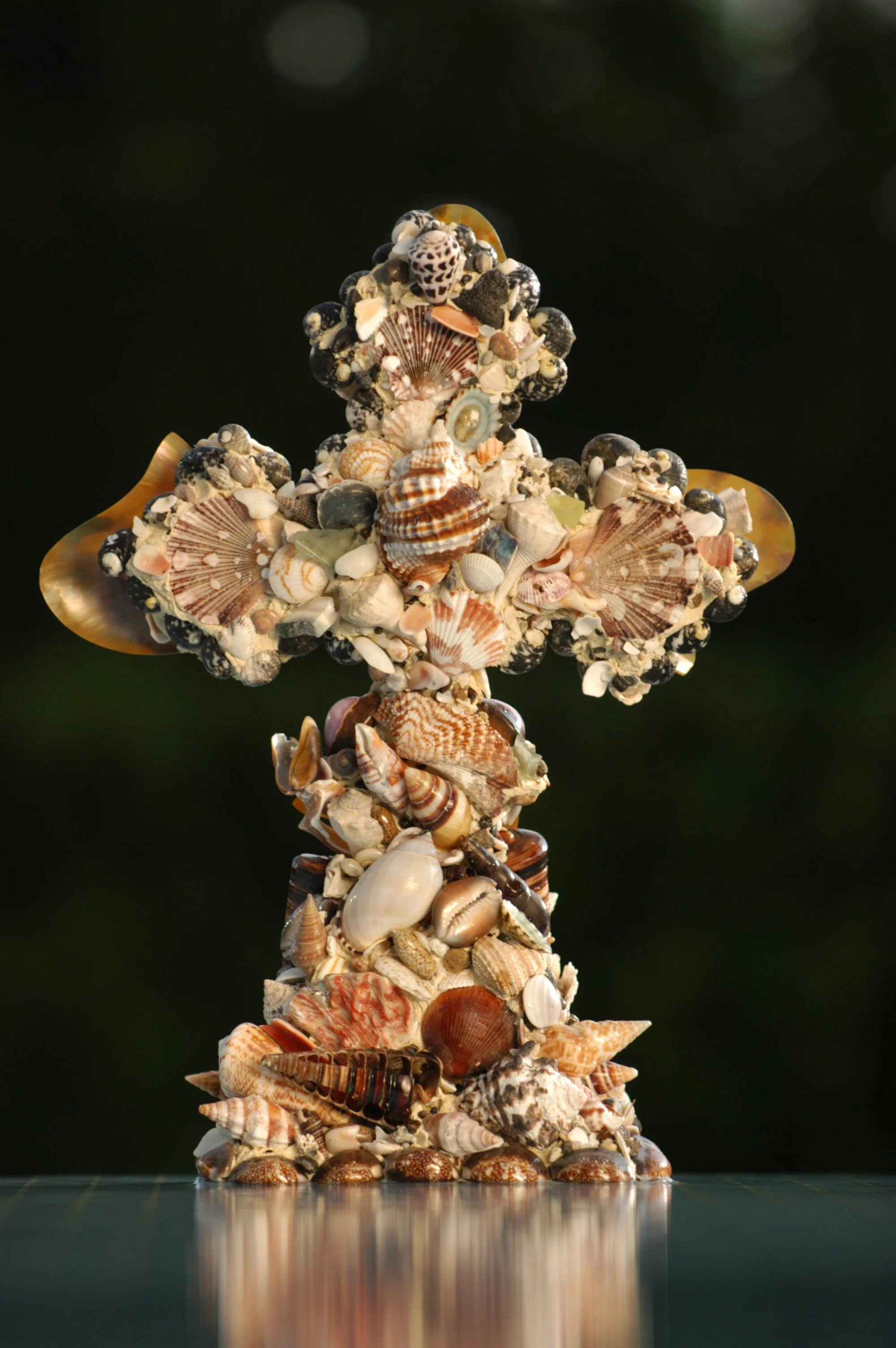 A tall sculpture made entirely of various seashells arranged in a layered and intricate pattern, forming a conical shape against a dark background.