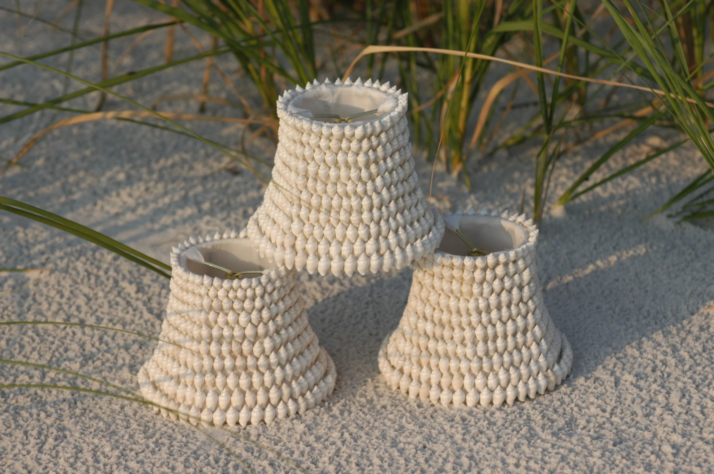 Three white woven lanterns with cone-shaped design placed on white sandy beach with green beach grass in the background.
