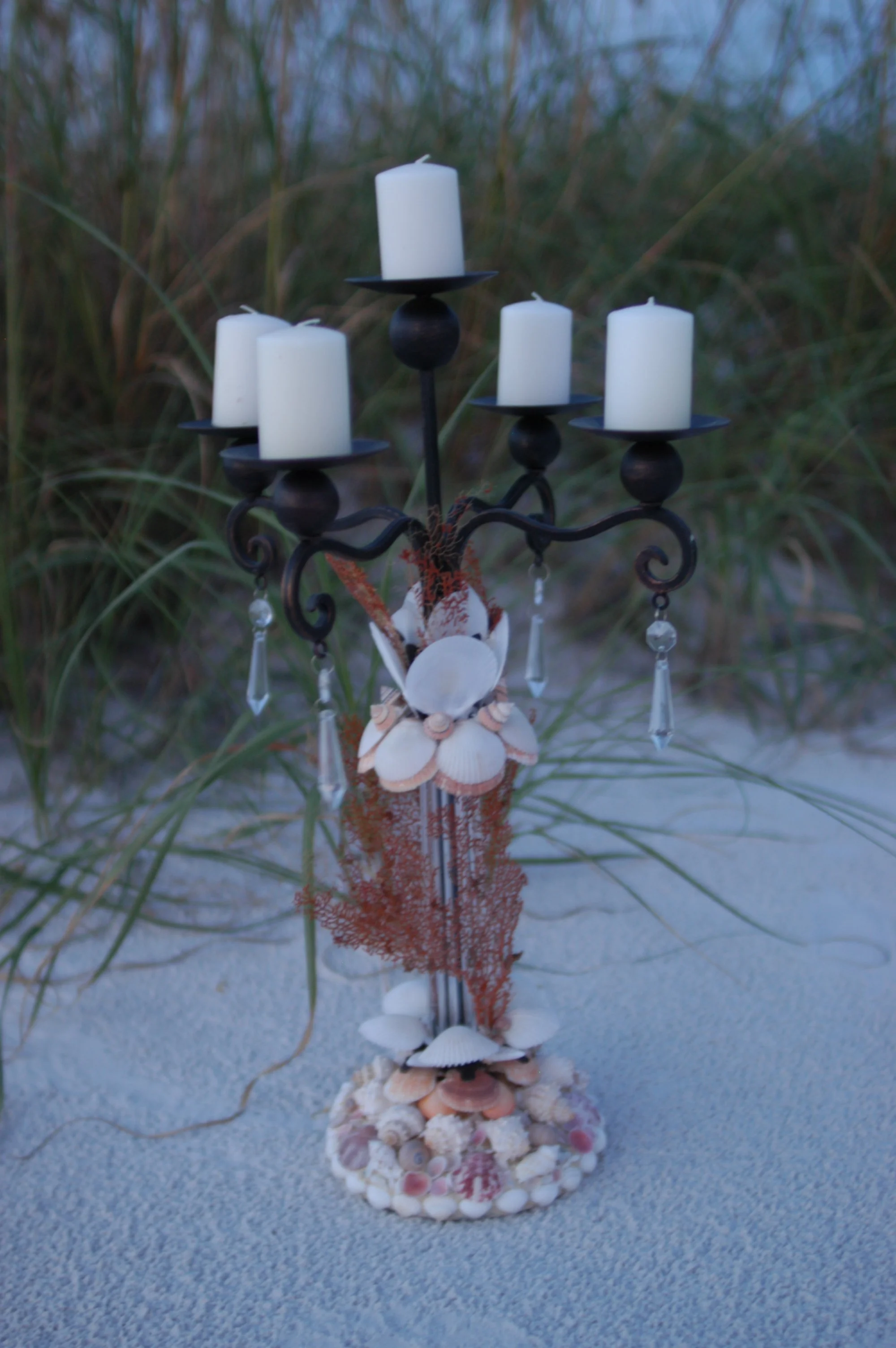 A decorative candelabra adorned with seashells, hanging crystals, coral, and other beach-themed ornaments, arranged on a sandy surface with sparse grass in the background.