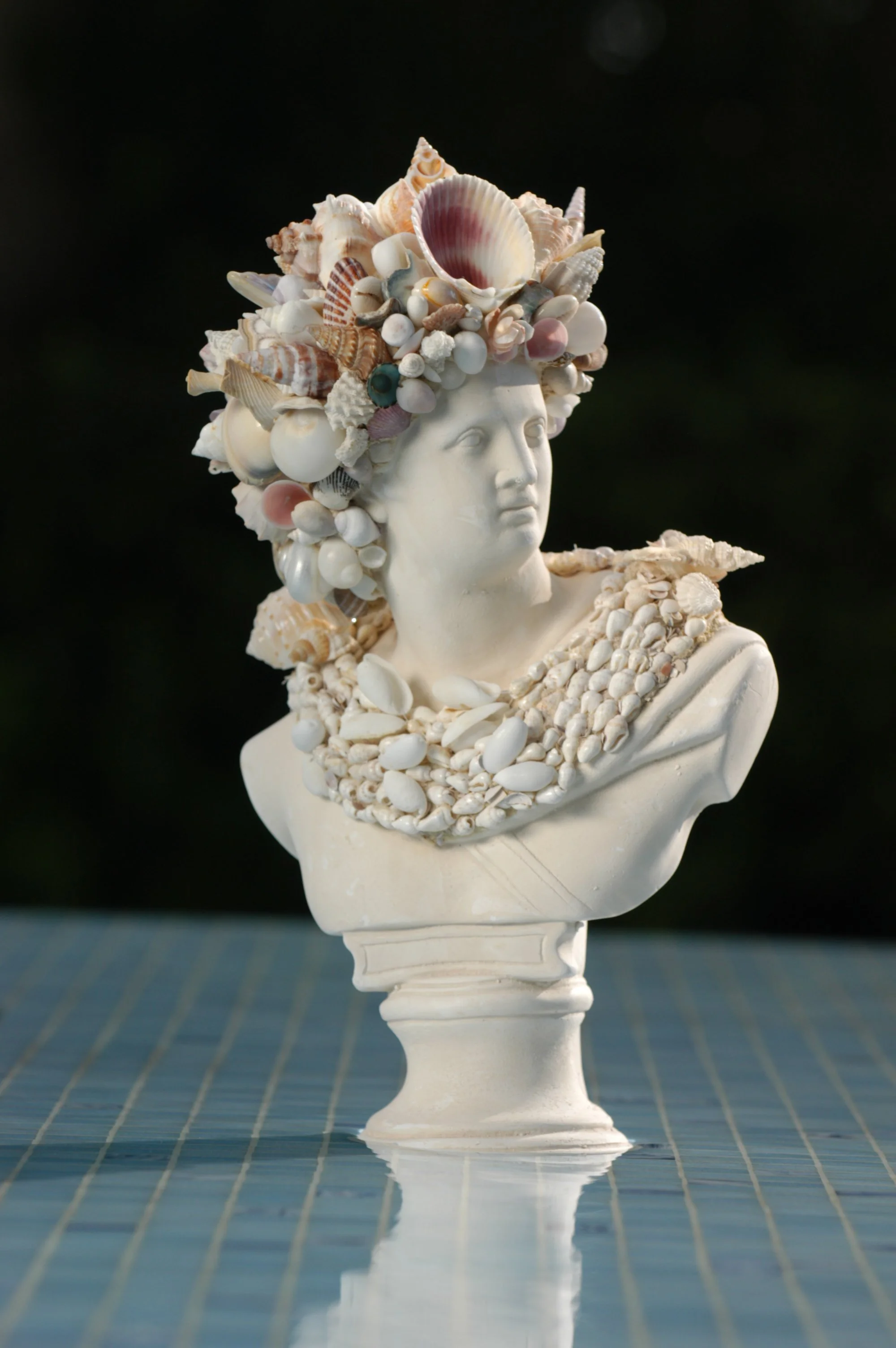 A bust sculpture of a woman adorned with an elaborate headdress made of various seashells, placed on a white pedestal.