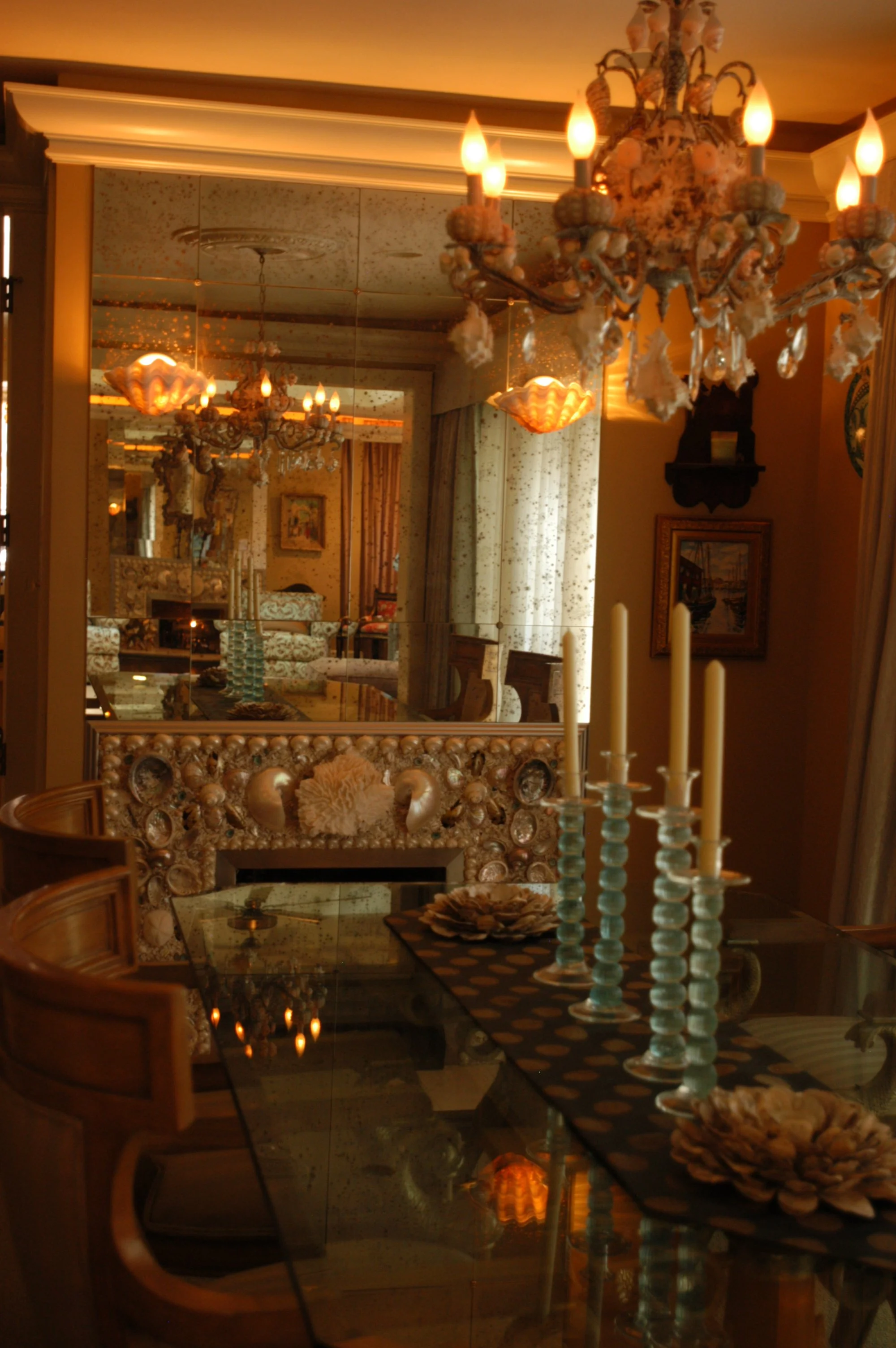An interior room with a reflection of a chandelier, a mirror, and various decorative items including candles, a painting, and floral arrangements.