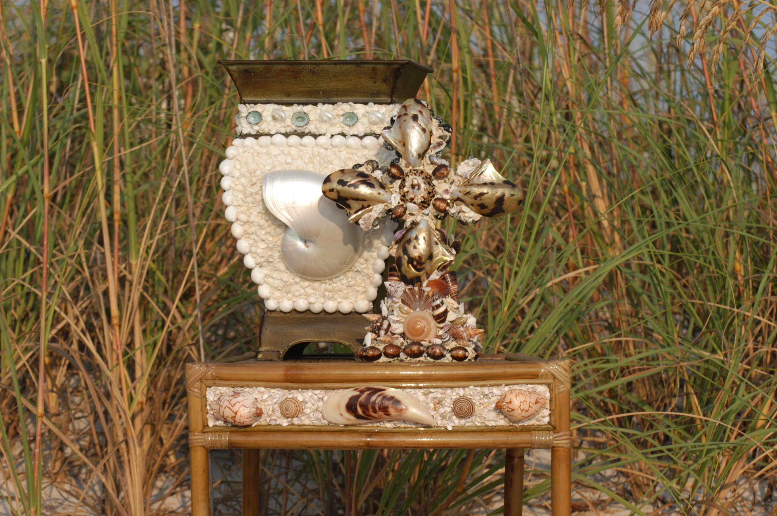 Decorative display of seashells, pearls, and a tree frog on a small wooden stand outdoors, with tall grass in the background.