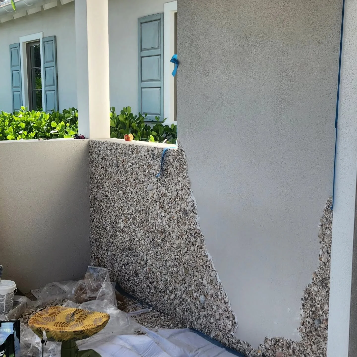 Under construction or renovation space with unfinished wall and granite slab, construction tools and supplies, and a partially built outdoor balcony with green shrubbery and windows in the background.
