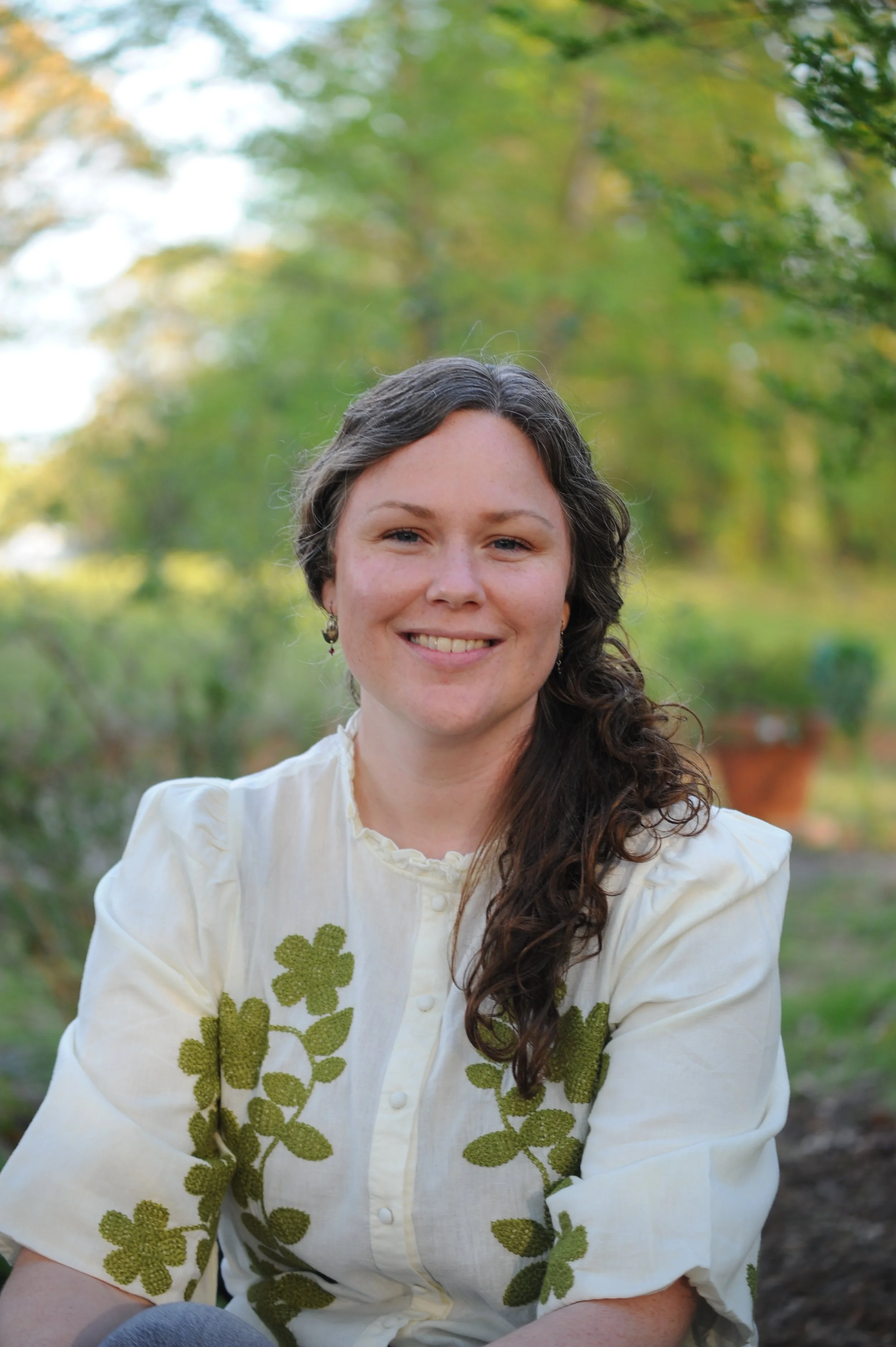 A woman with long, curly brown hair wearing a white blouse with green embroidery, smiling, outdoors with green trees and plants in the background.