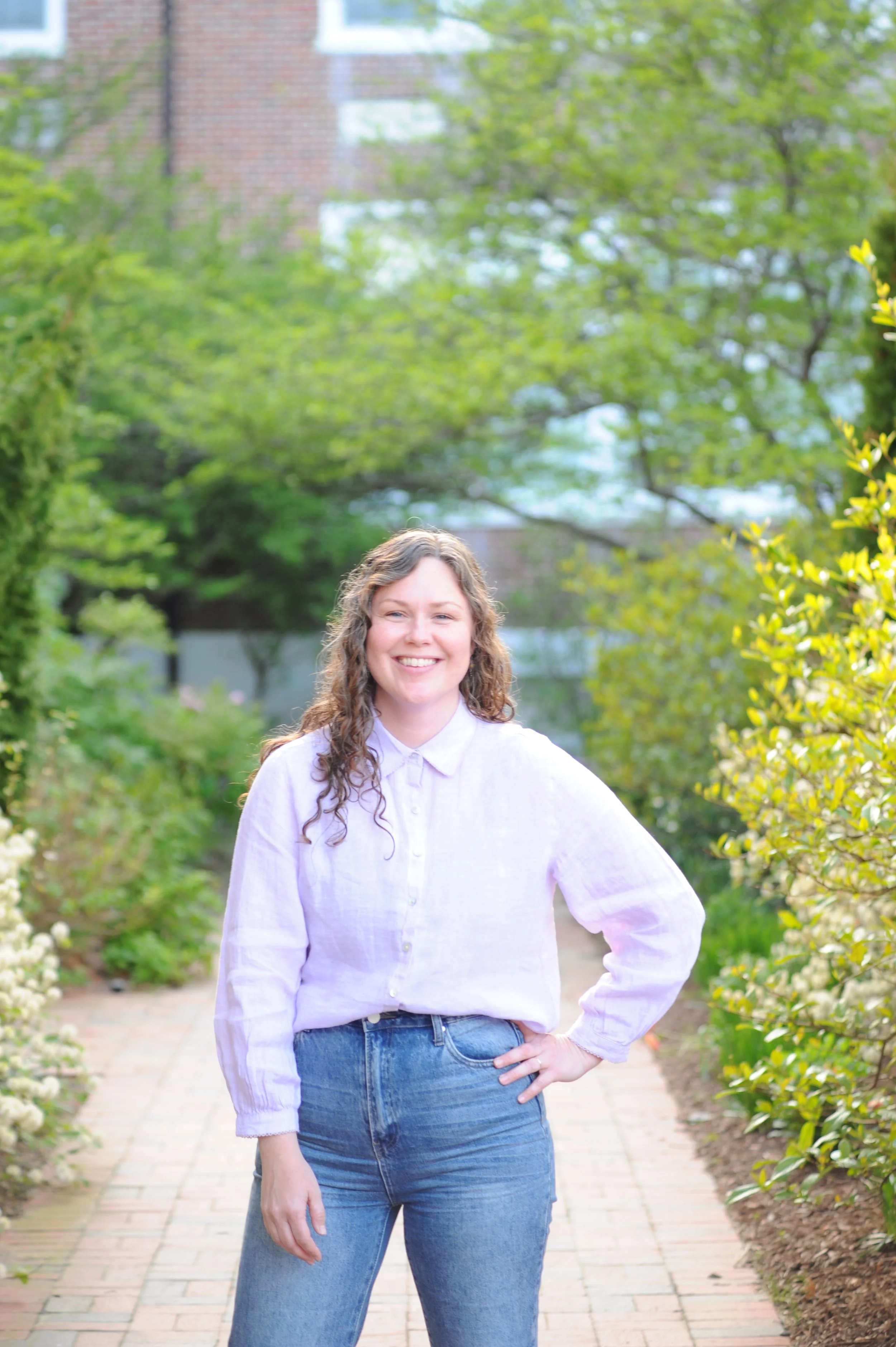 A young woman with curly hair wearing a white button-up shirt and blue jeans standing on a garden path with green trees and bushes surrounding her.