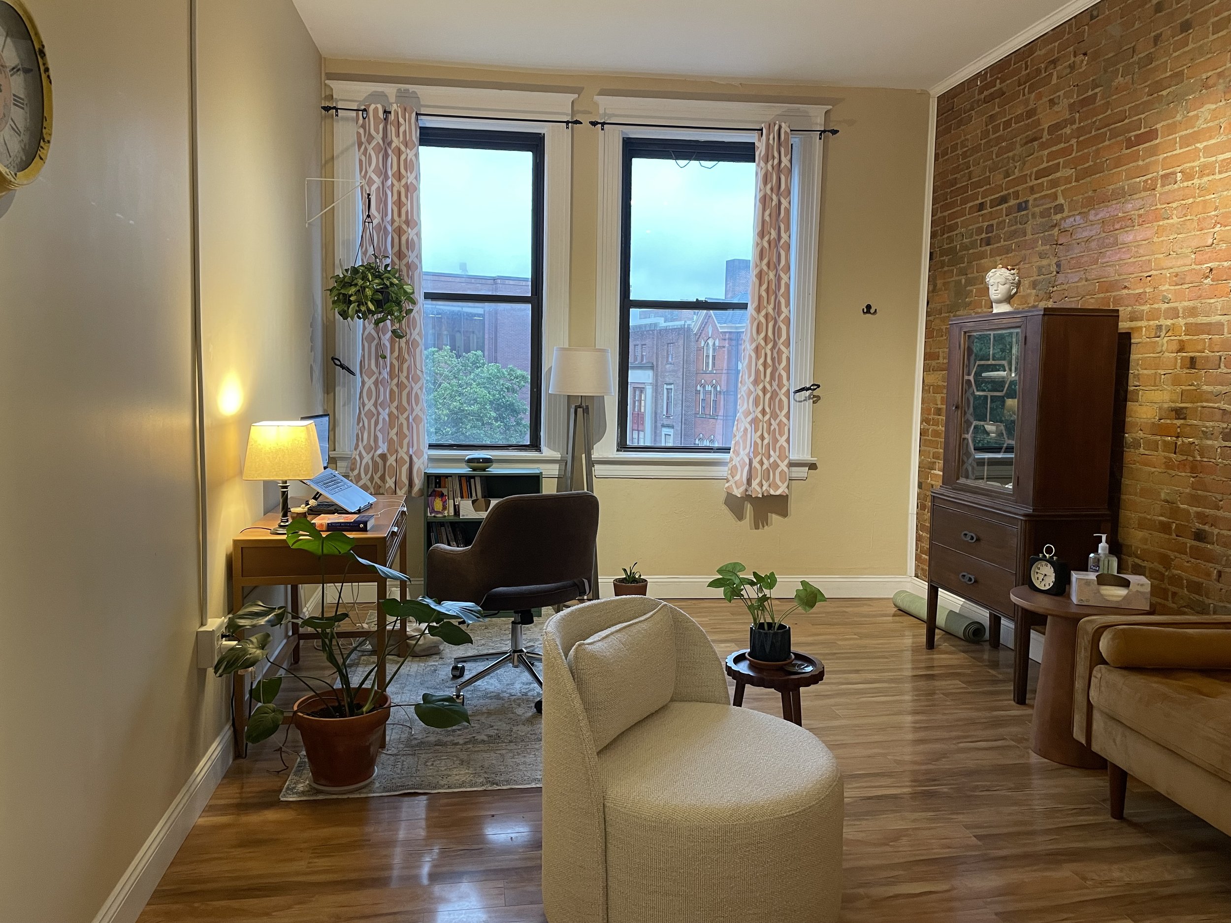 A cozy living room with a large brick wall and two large windows covered by patterned curtains. There is a small wooden cabinet with decorative items, including a bust sculpture. The room features plants, a floor lamp, a chair, and a couch, with hardwood floors and warm lighting.