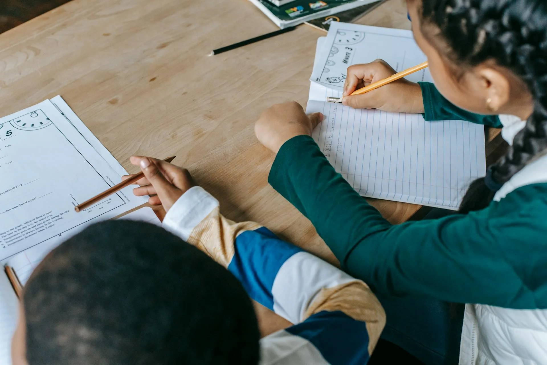 Two children working on math worksheets at a wooden table, writing in notebooks with pencils.