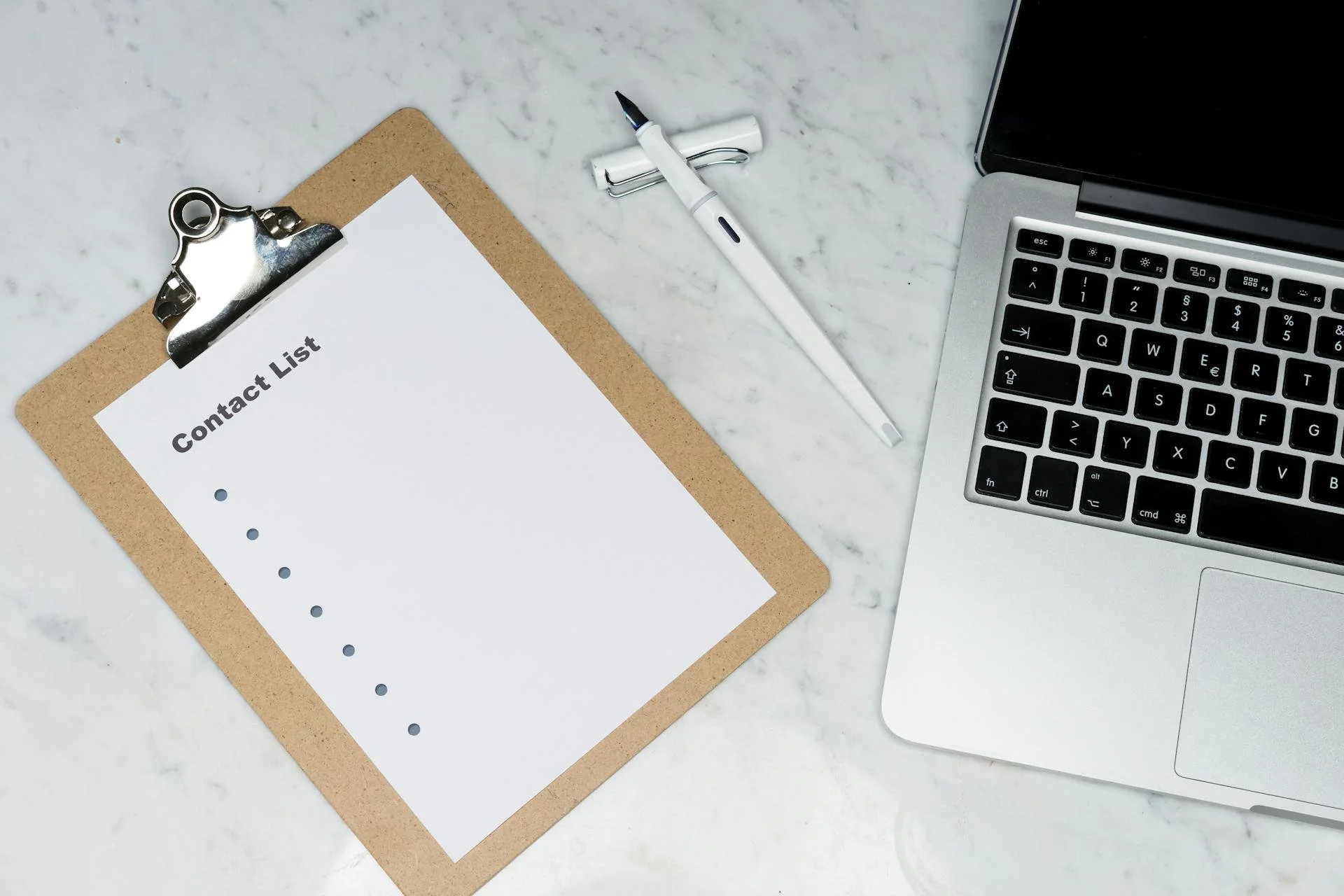 A clipboard with a contact list on white paper, a white pen, a laptop, and a marble surface.