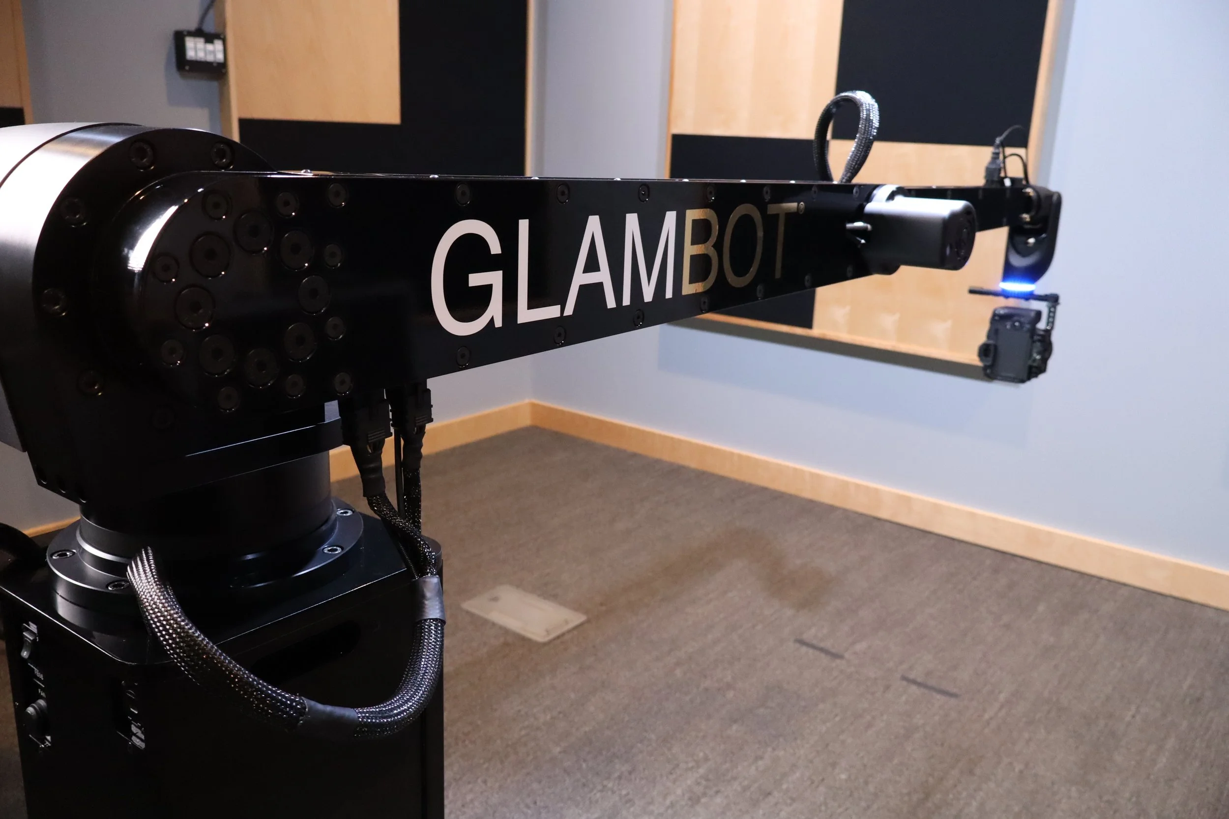 A robotic arm with the label 'GLAMBOT' printed on it, situated indoors on a carpeted floor, with soundproofing panels on the wall in the background.