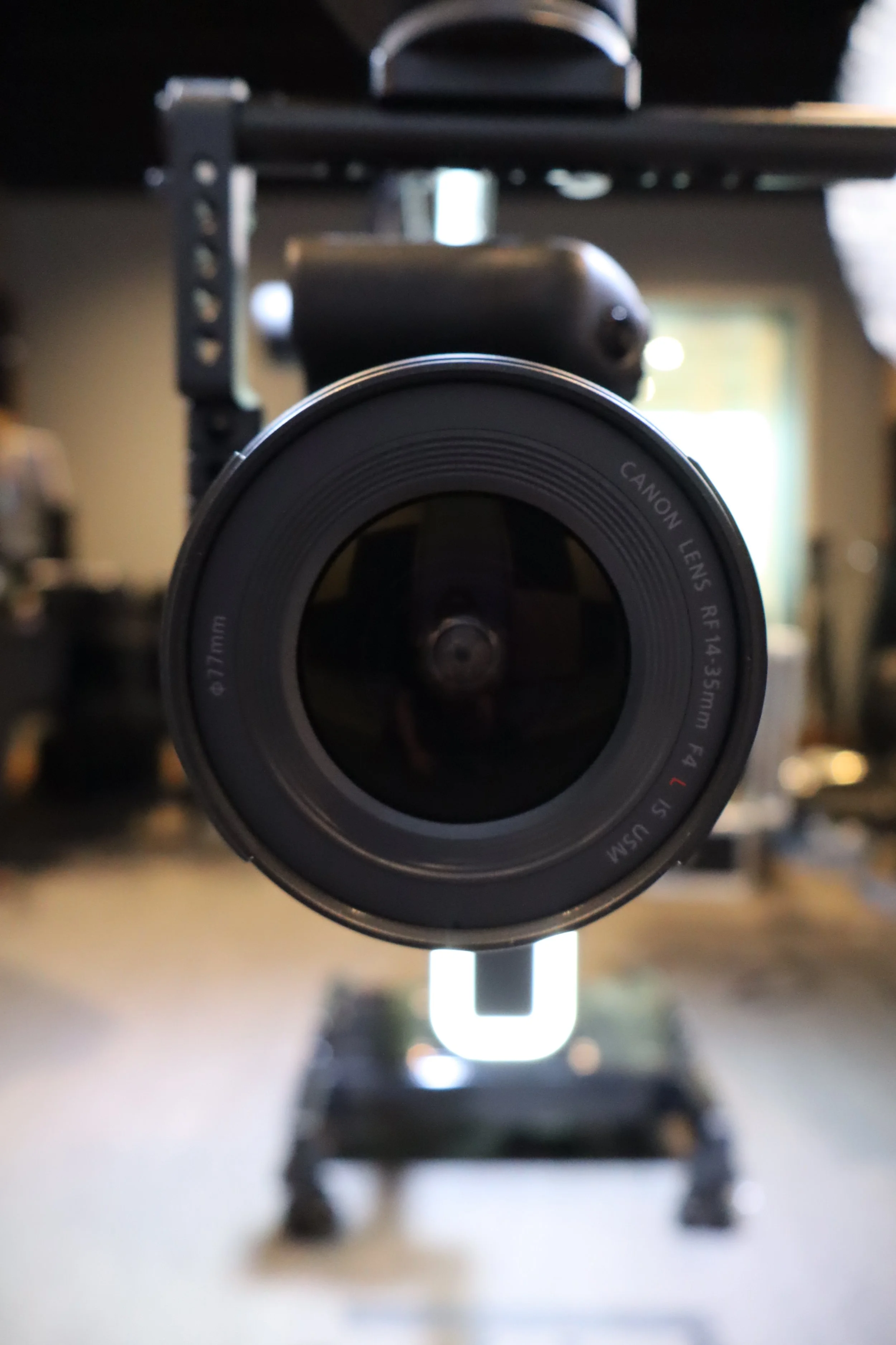 Close-up of a camera lens in focus, mounted on a camera body with a blurred background of a room with shelves and a person.