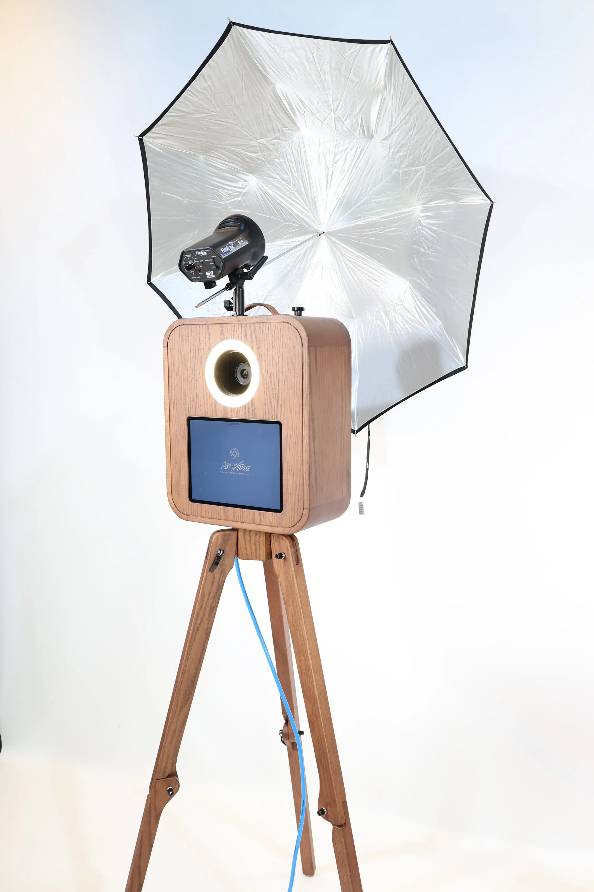 Photo of a photo booth with a wooden case, mounted on a wooden tripod. The booth includes a camera, a logo on the front, and a ring light. It is set up with a large white umbrella light reflector positioned above it for lighting.