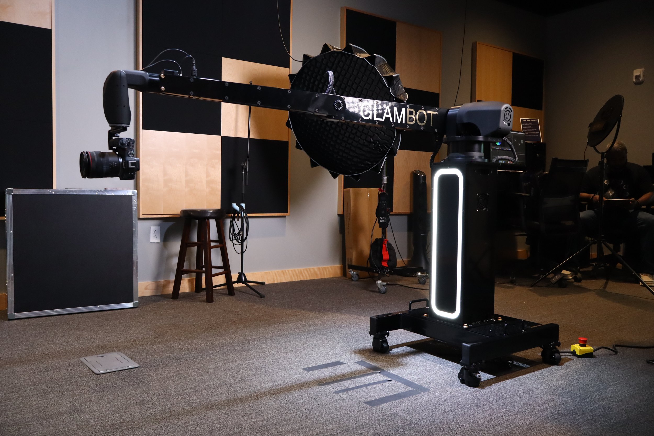 A robotic arm labeled 'GLAMBOT' in a room with acoustic panels, a stool, a camera mounted on the robotic arm, and various electronic equipment.