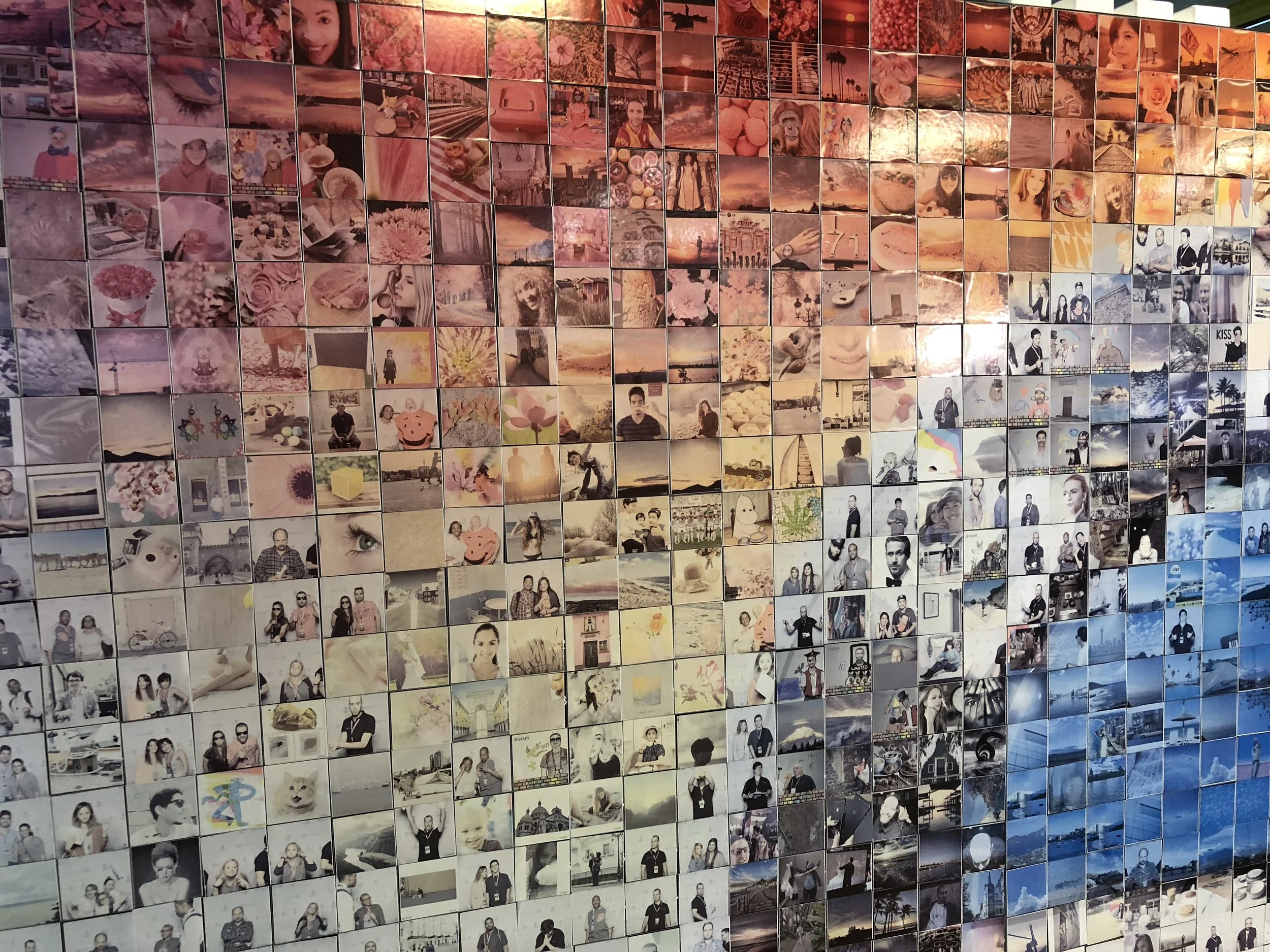 A large wall covered with many small photos arranged in a grid pattern, showcasing various landscapes, portraits, objects, and scenes in warm pinkish tones.