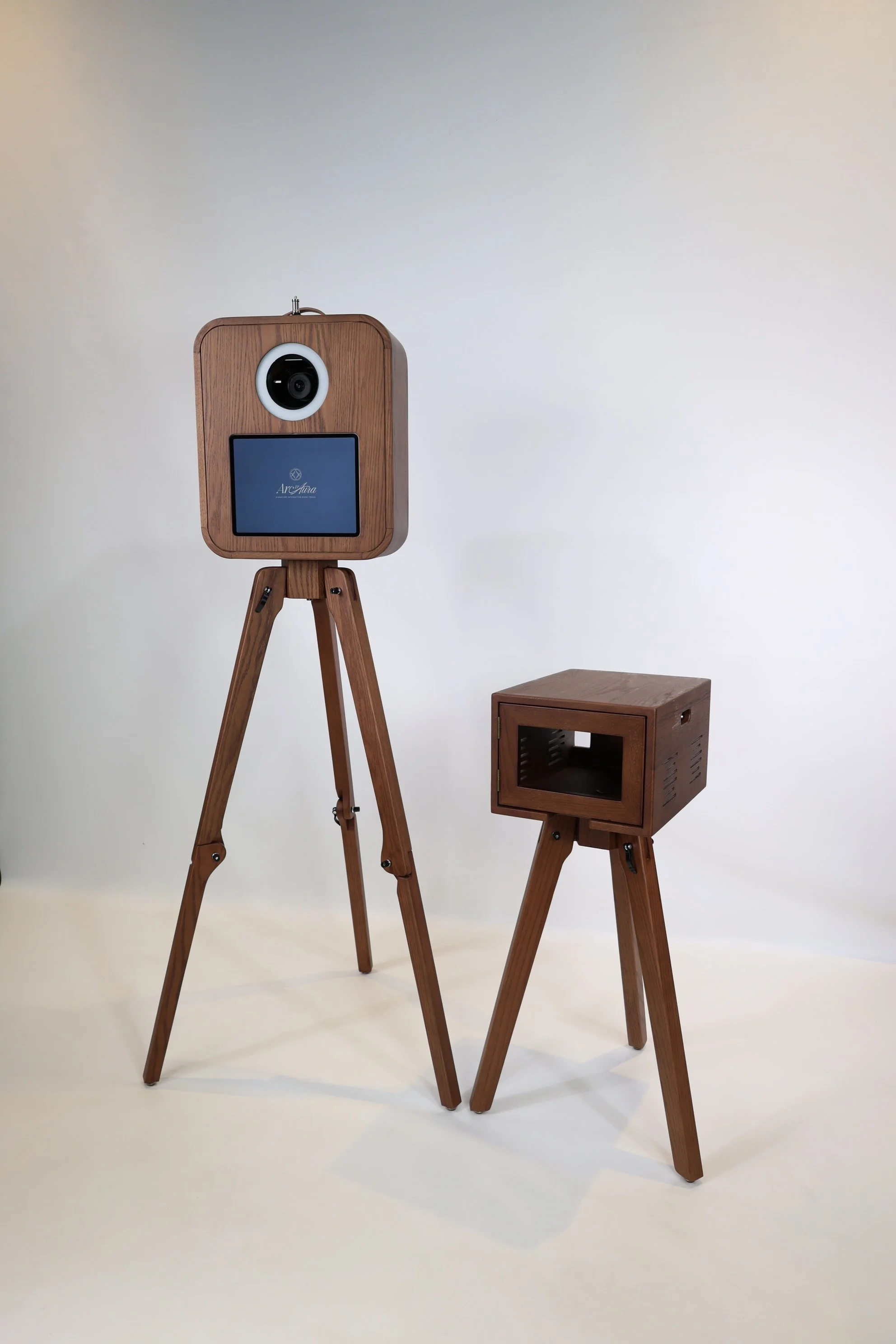 Two wooden camera-like structures on wooden tripods with rectangular bodies, one with a screen displaying 'ArcAura' and the other with a circular opening for a camera lens, placed against a plain white backdrop.