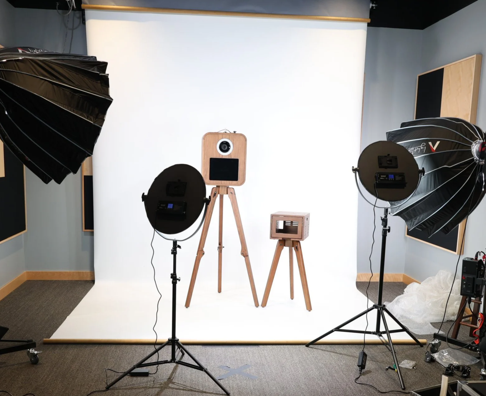 Photography studio setup with a white seamless backdrop, two large studio lights with umbrellas, a wooden camera on a tripod, and other photography equipment.
