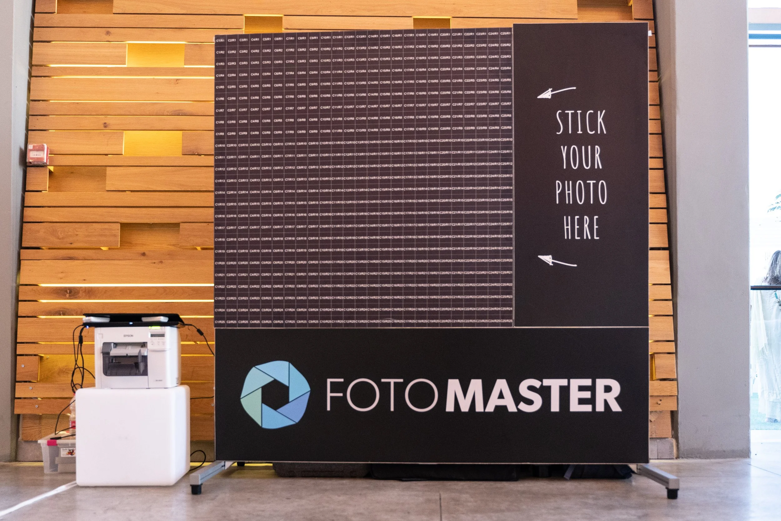 Photo booth setup with a wooden backdrop and a large black sign that says 'Stick Your Photo Here' with arrows pointing to the space for photos. A photocopier is visible in the foreground.