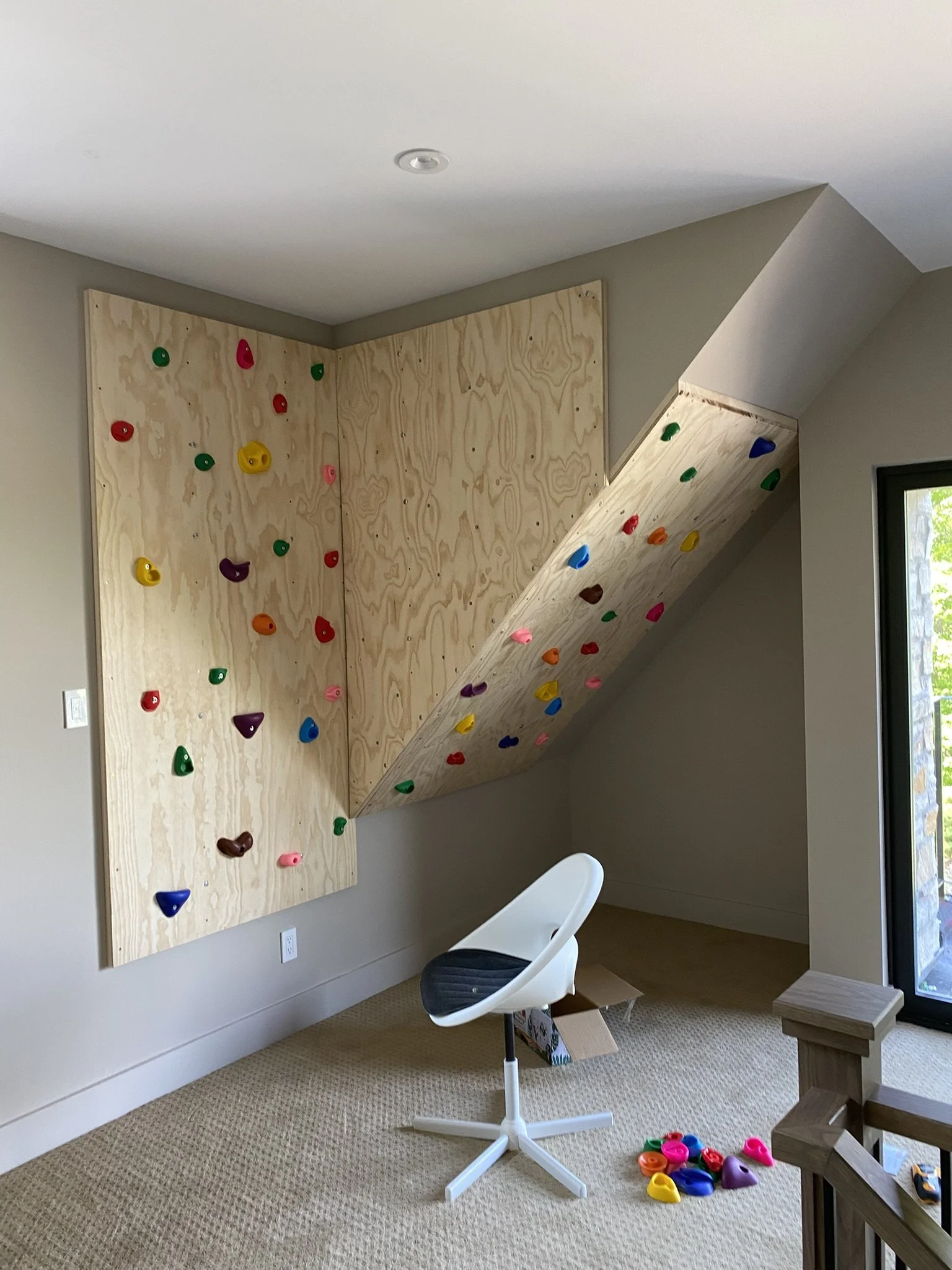 Indoor rock climbing wall with colorful holds, built into a corner, with a beige carpeted floor and a modern white chair nearby.