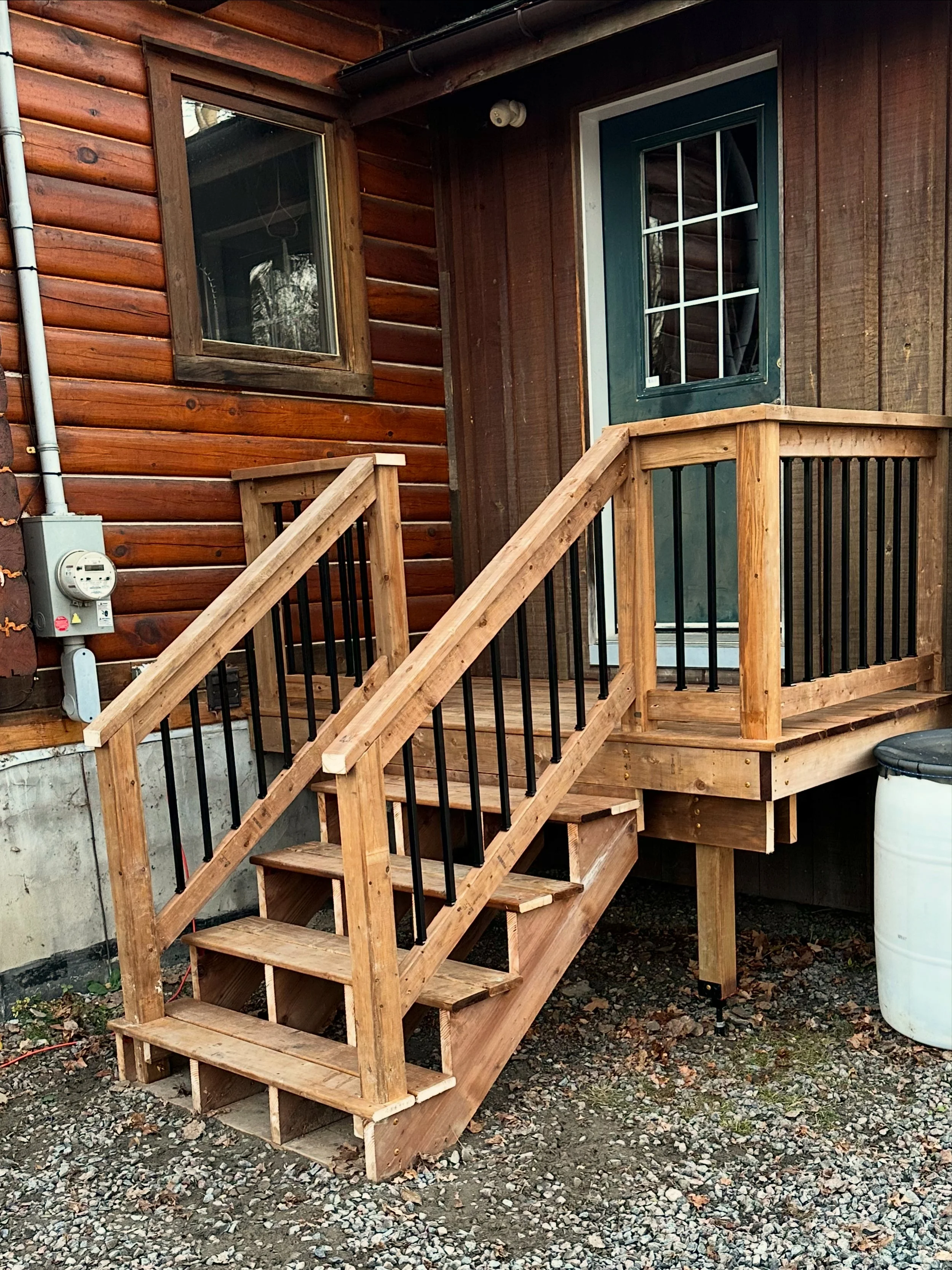Wooden staircase leading up to a small porch with railings, attached to a house with wood siding and a glass panel door.