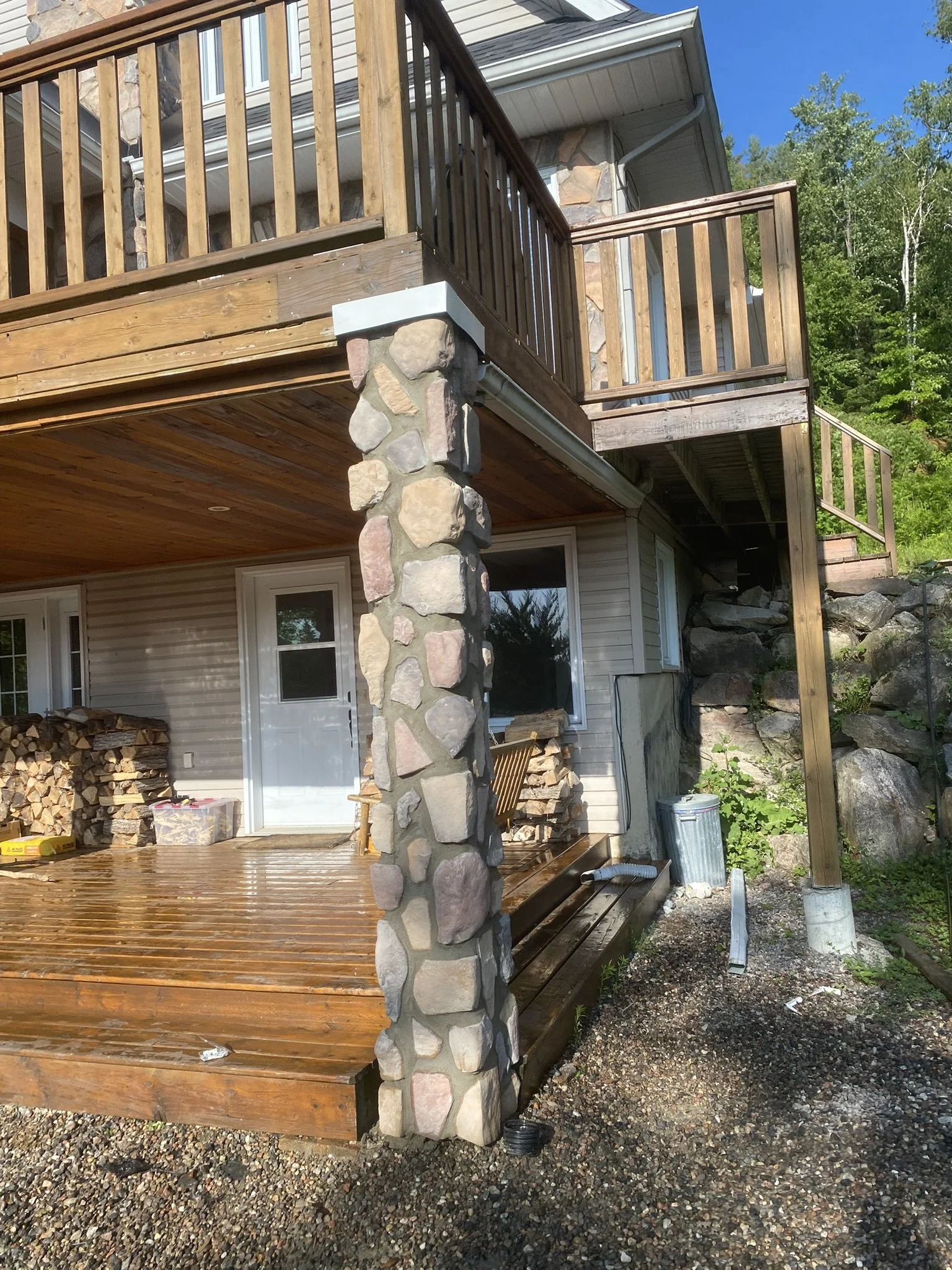 The photo shows the back of a house with a new wooden deck and railing on the upper level, supported by a stone pillar. There is a waterproofed and polished lower deck area, with firewood stacked against the house wall. Part of the yard with gravel and a trash can is visible, and trees are in the background under a clear blue sky.