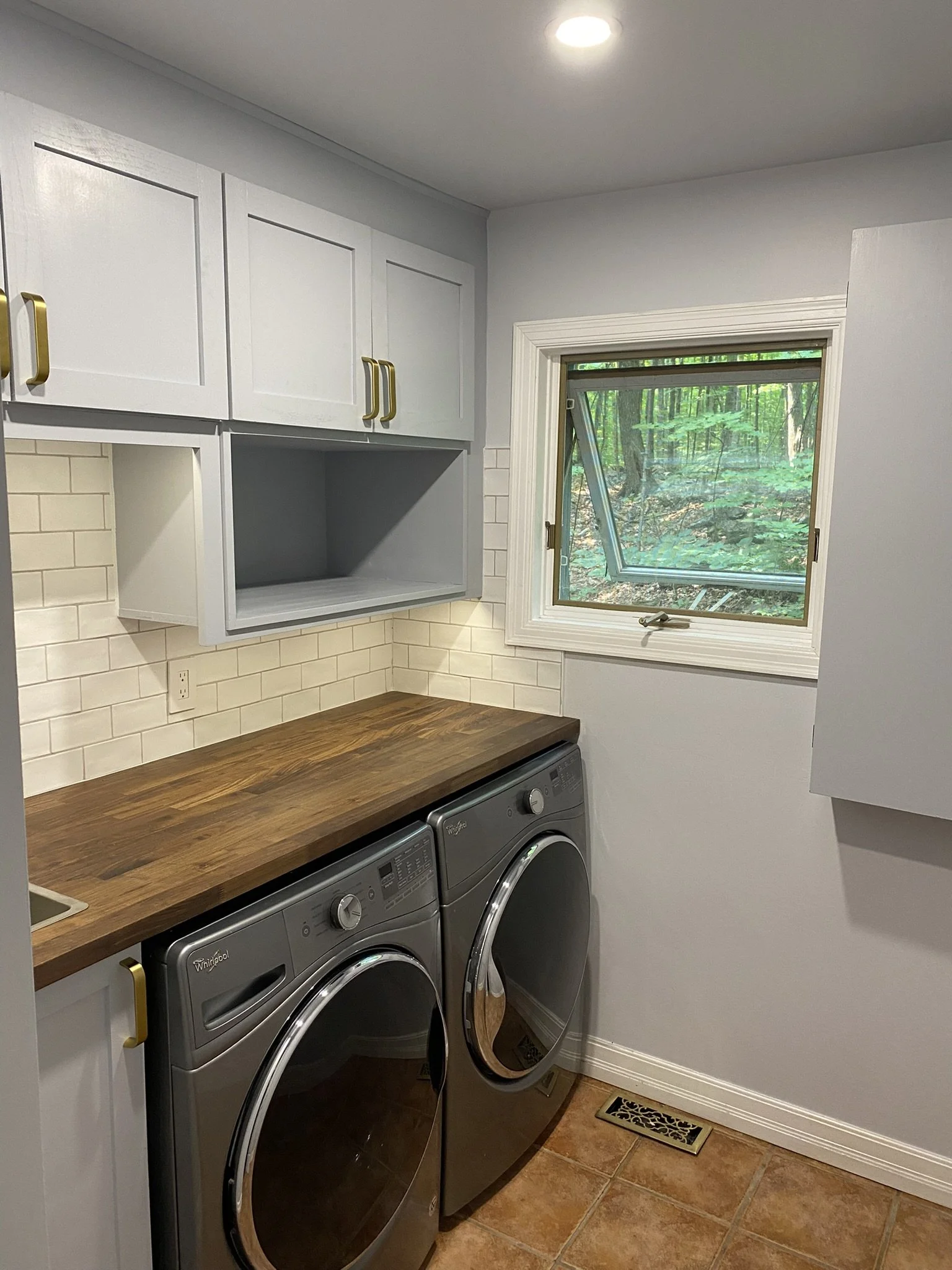 Laundry room with white cabinets, wooden countertop, front-loading washer and dryer, window showing trees outside, beige tile floor, white brick backsplash.