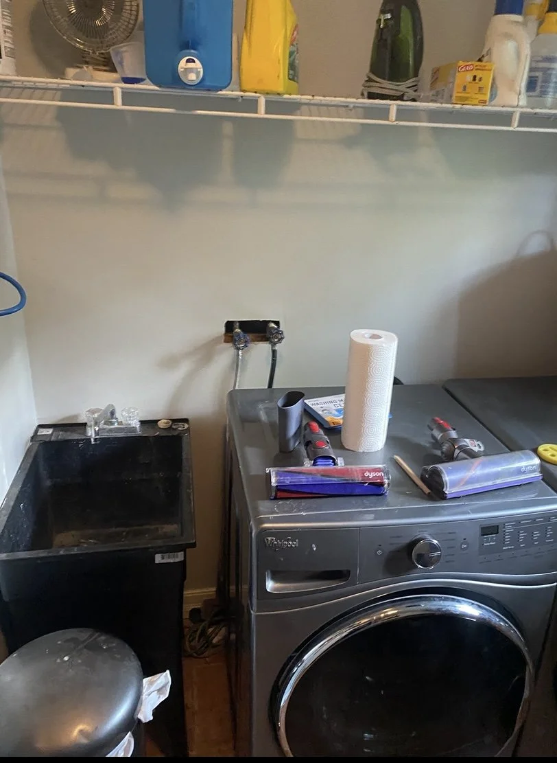 Laundry room with white wire shelf holding detergent, cleaning supplies, and bottles; washing machine with paper towel roll, tools, and cleaning products on top; utility sink, trash can, and a fan visible in the space.