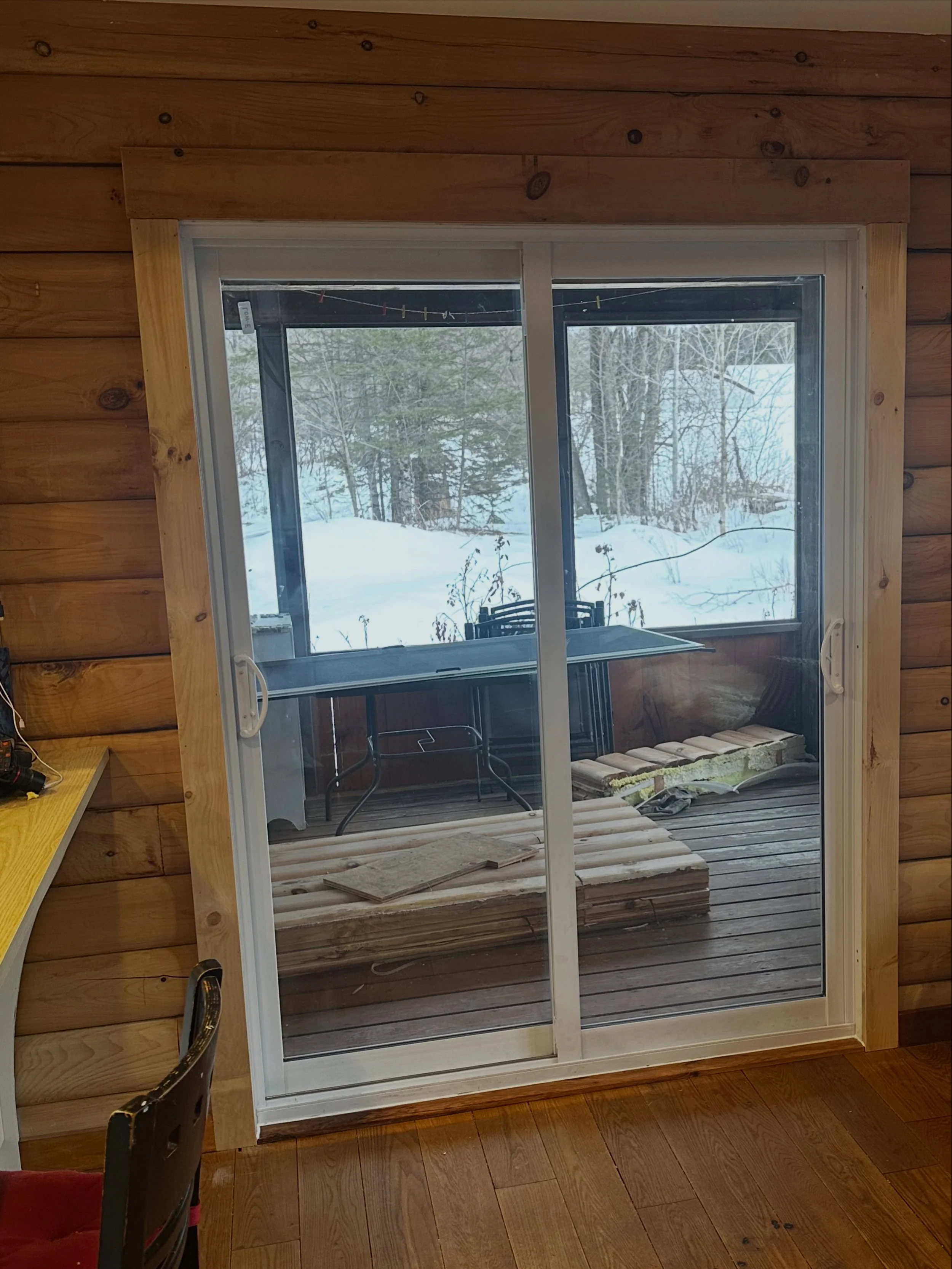 View through a sliding glass door showing a snowy outdoor scene with bare trees, a wooden deck, construction wood, and outdoor furniture.