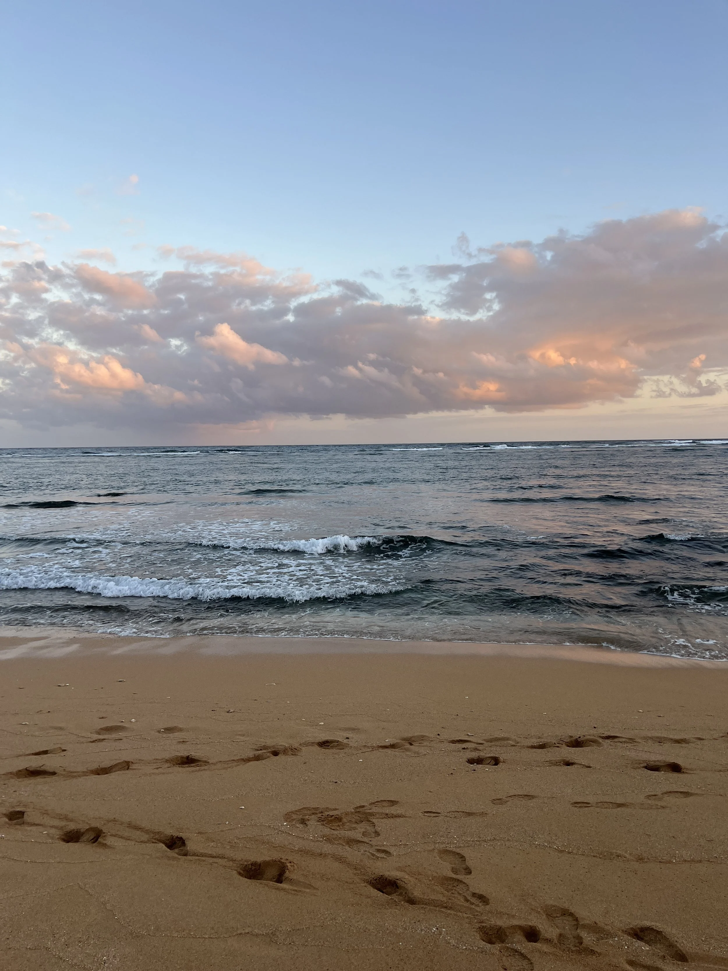 A sandy beach with footprints walking next to the ocean. The sky is partly cloudy with hints of pink and orange, indicating the time is either sunrise or sunset. Gentle waves are visible on the water.