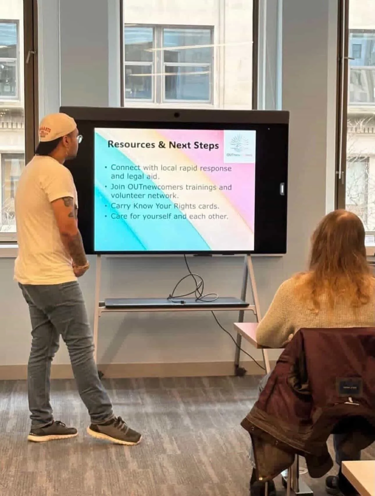 A person in a white T-shirt and gray jeans is standing and looking at a presentation slide on a large screen, while another person with red hair seated nearby listens. The slide shows resources and next steps.