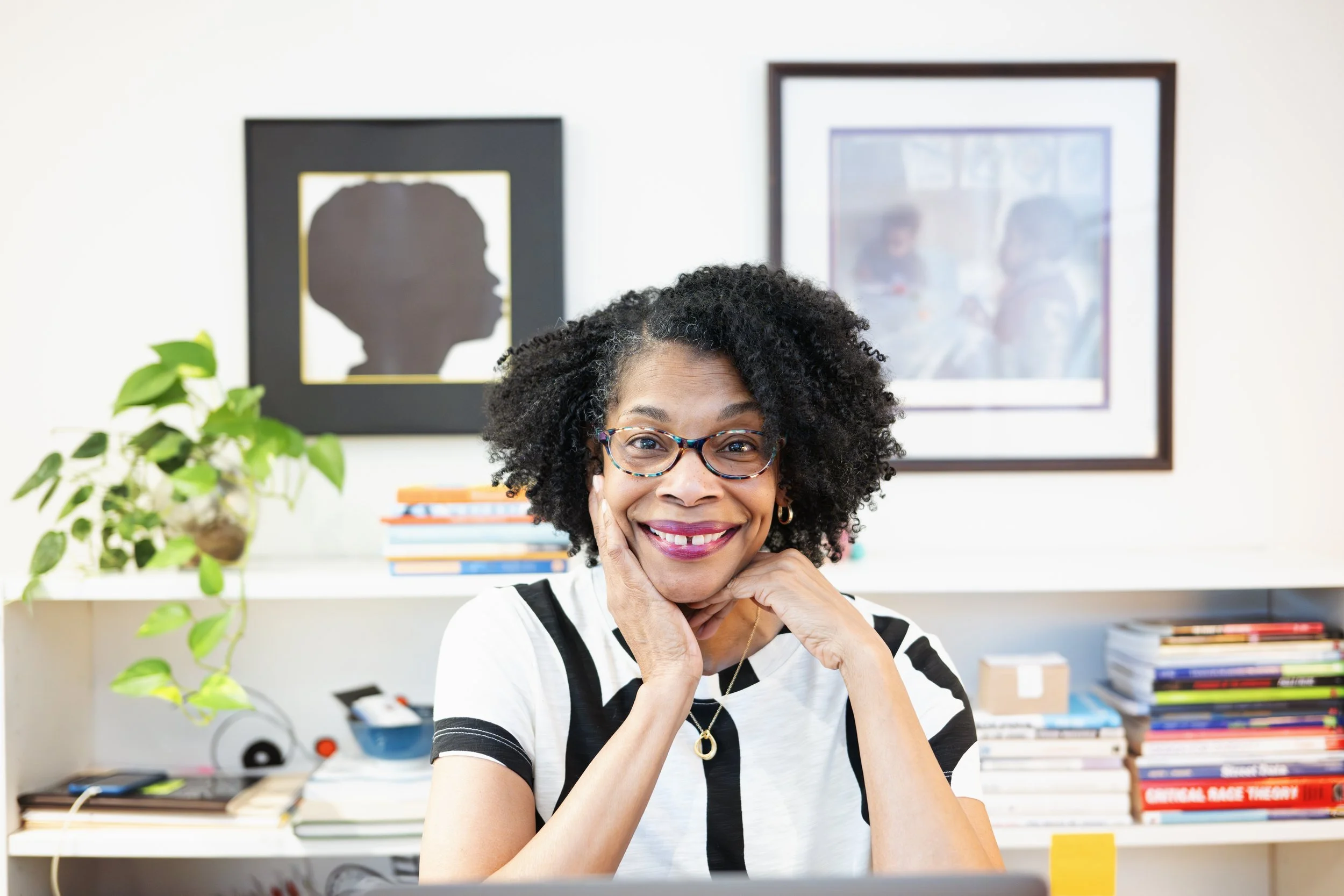 A woman with curly hair and glasses smiling, resting her chin on her hand in an office or home workspace with framed artwork behind her and a plant on her left.