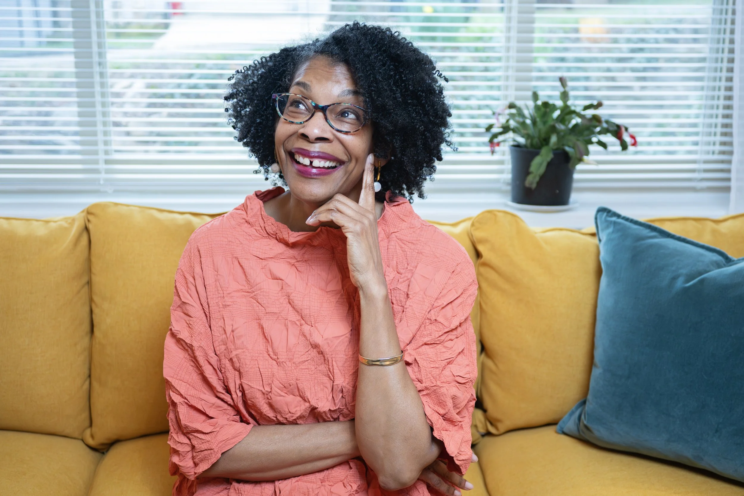 A woman with black curly hair and glasses, wearing a coral blouse, sitting on a yellow couch, smiling, with her hand against her cheek, in front of a window with blinds and a houseplant in the background.